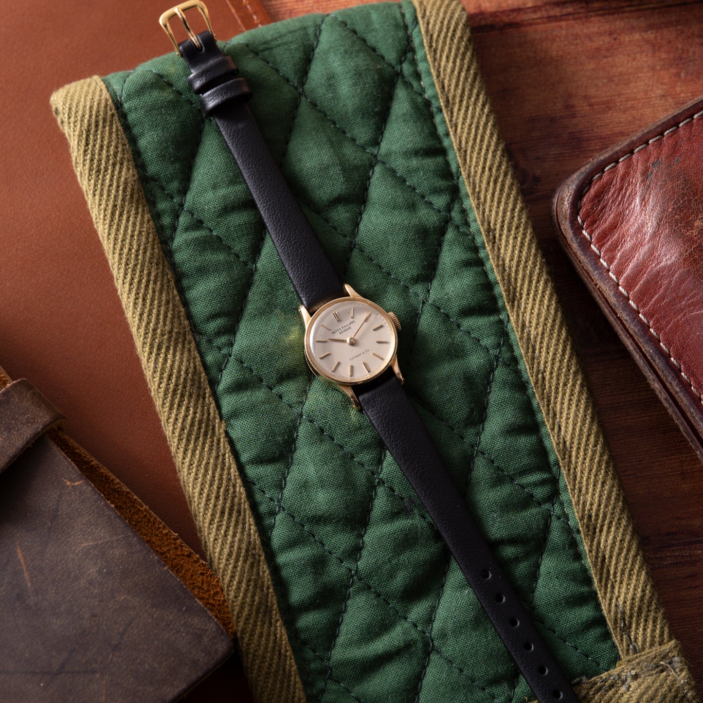 A Patek Philippe Calatrava 'Tiffany & Co' ladies dress watch in 18K yellow gold with a black leather strap sits on a green quilted fabric roll, surrounded by brown leather accessories on a wooden surface.
