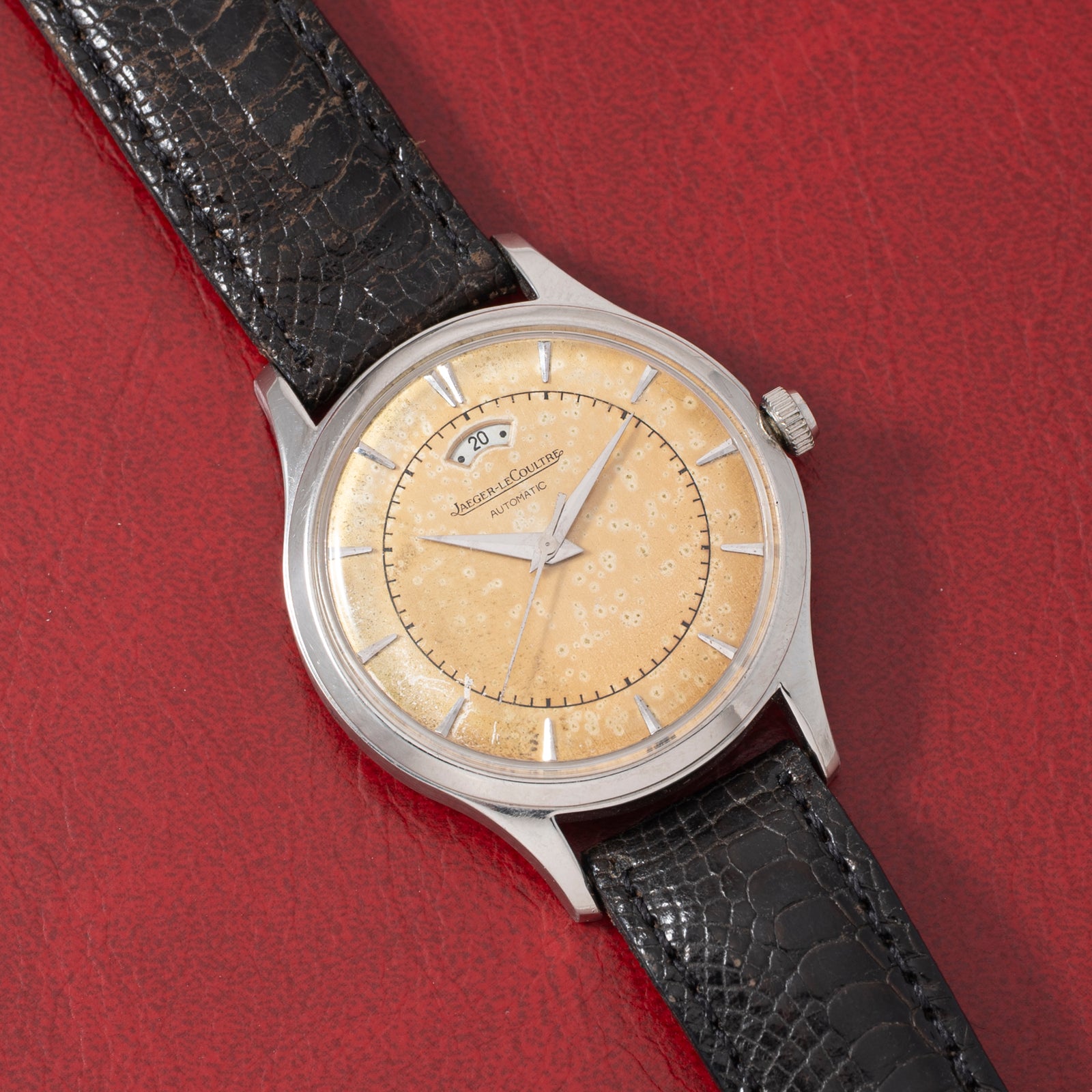 A vintage Jaeger LeCoultre 'European Market' Powermatic dress watch with a textured beige dial, silver hour markers, power-reserve display, date window, and black leather strap displayed on a red textured surface.