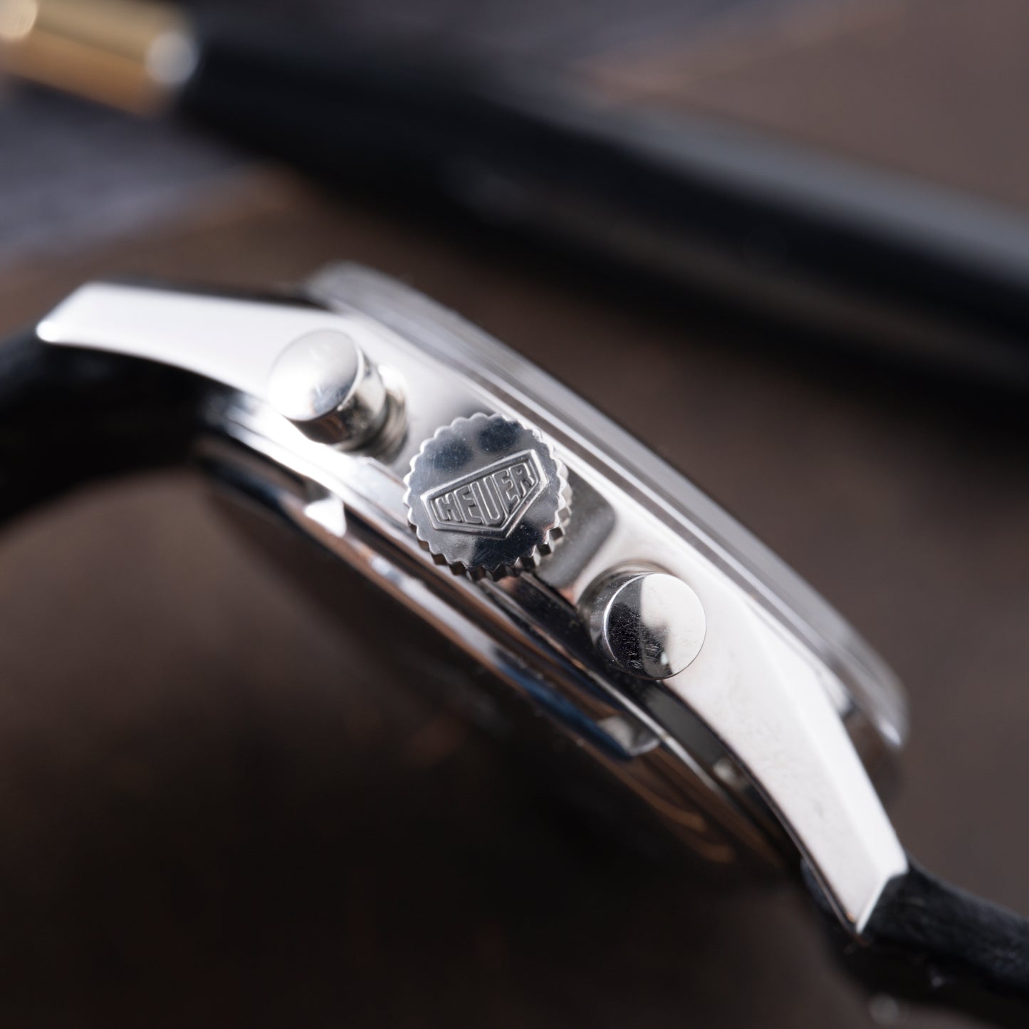 Close-up side view of a TAG Heuer 1964 Carrera Re-Edition, highlighting the metallic crown engraved with TAG Heuer and two pushers. The watch features a black strap, with a blurred pen visible in the background.