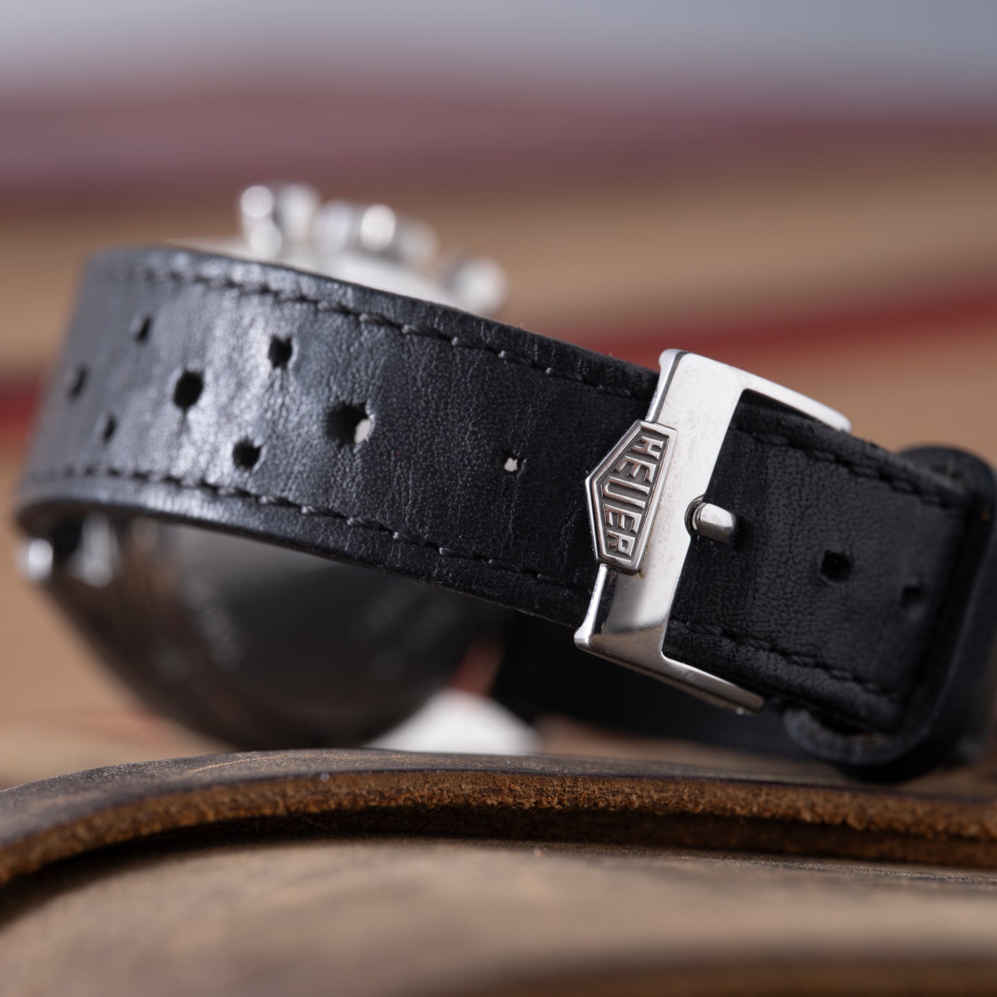 Close-up of a black leather strap with multiple holes and a silver buckle engraved "Heuer." In the background, the blurred dial of the TAG Heuer 1964 Carrera Re-Edition rests on a brown leather surface.