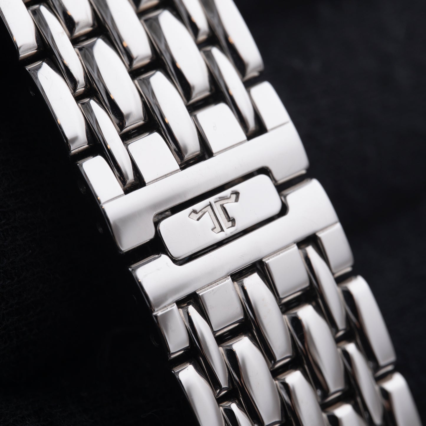 Close-up of a polished silver watch band with interlocking links and a clasp engraved with a stylized T logo, set against black—an elegant match for the Jaeger LeCoultre Master Control Chronograph Mecaquartz.