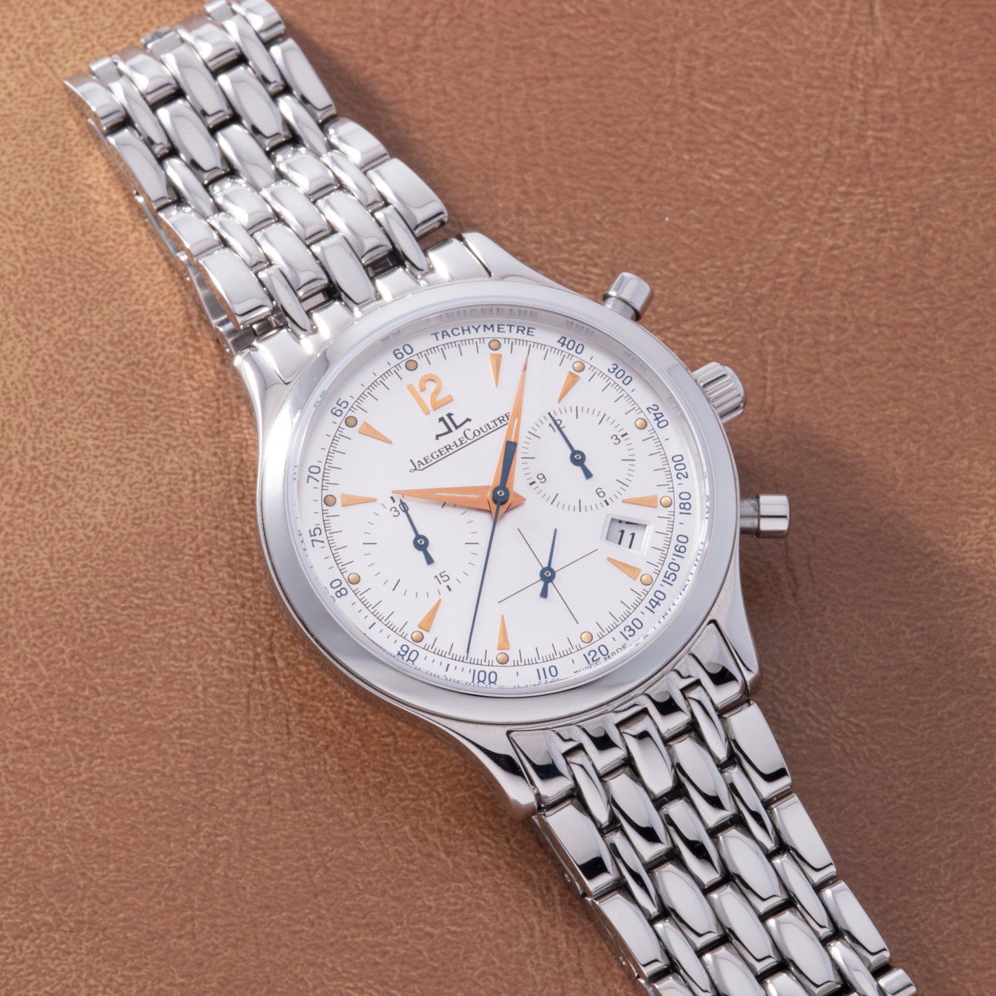 A Jaeger LeCoultre Master Control Chronograph Mecaquartz wristwatch featuring a white dial, orange hands, date display, and silver bracelet is shown on a brown textured surface.