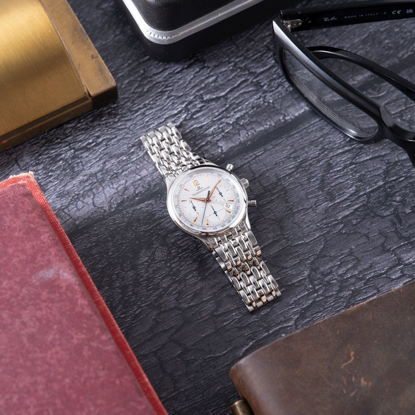A Jaeger LeCoultre Master Control Chronograph Mecaquartz with a silver case, white dial, and metal bracelet sits on a dark surface, surrounded by glasses, a red book, a brown leather accessory, and a brass item.