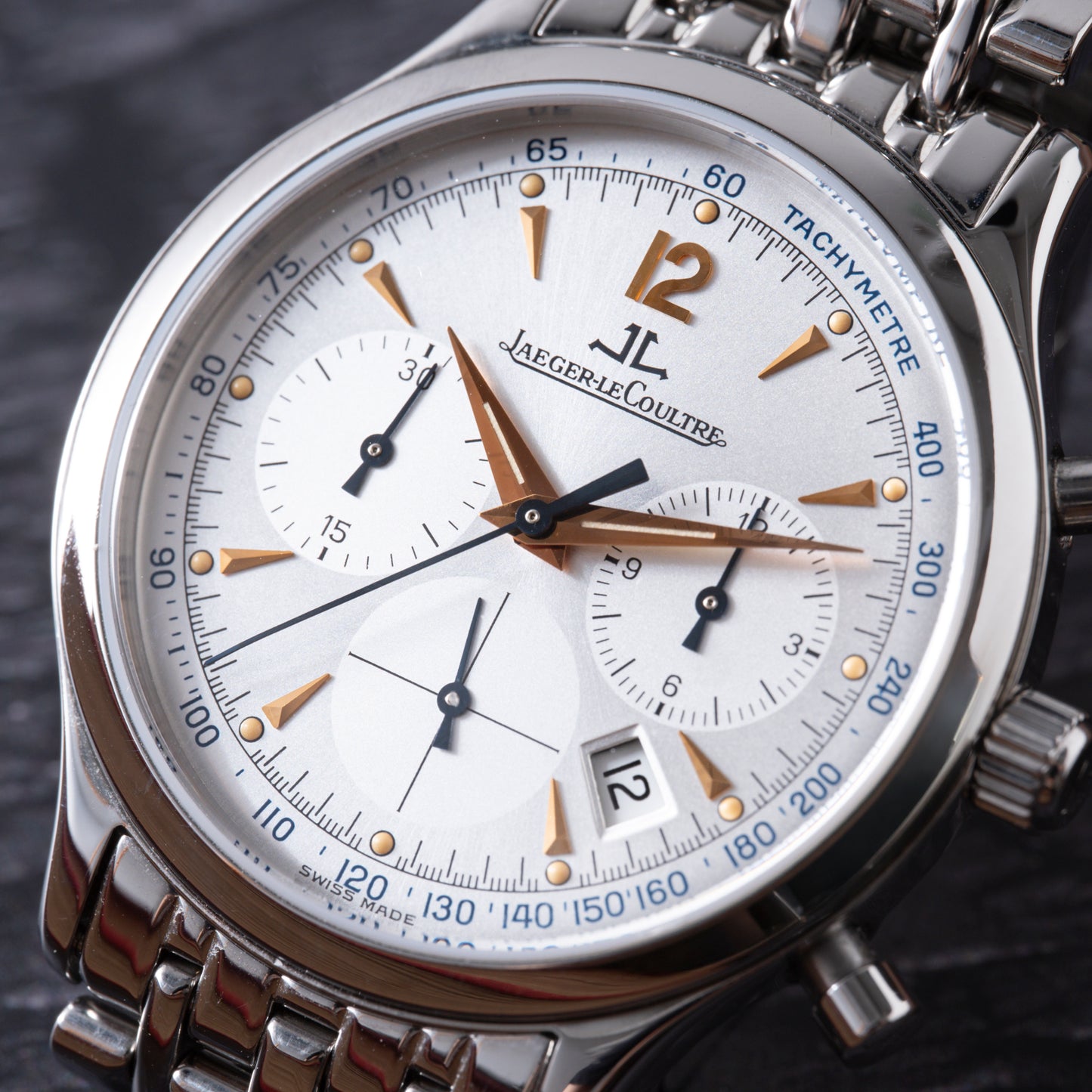 A close-up of the Jaeger-LeCoultre Master Control Chronograph Mecaquartz wristwatch by Jaeger LeCoultre, featuring a silver dial with gold accents, three subdials, date window, stainless steel bracelet, and detailed markings with the brand logo.