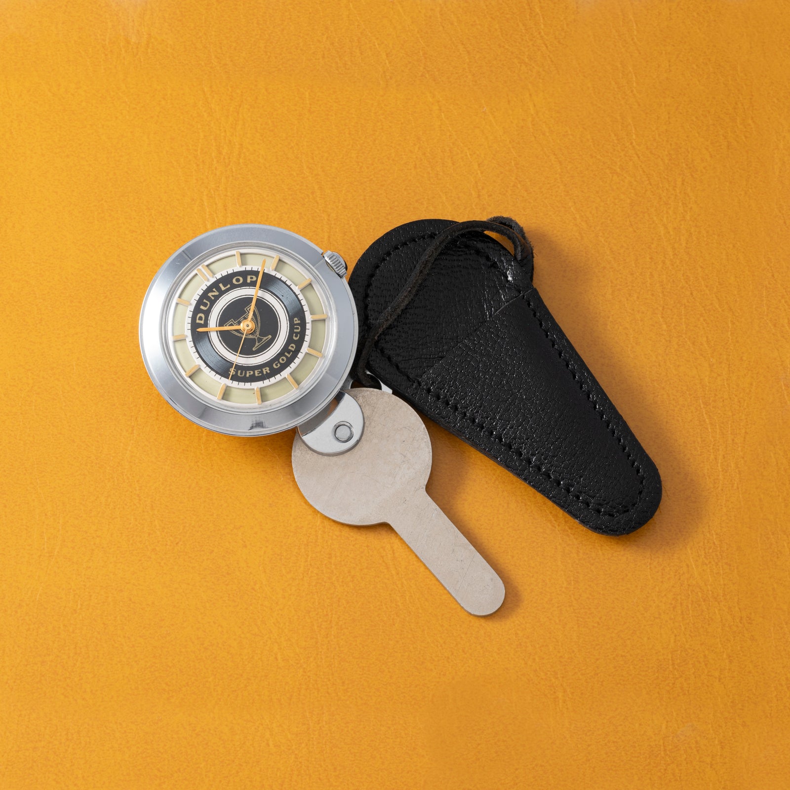 A vintage Prestige Watch Co. Dunlop Key Watch, featuring a round silver and black design on a beige key-shaped tag with a black leather case—perfect for classic car fans—rests on an orange leather surface.