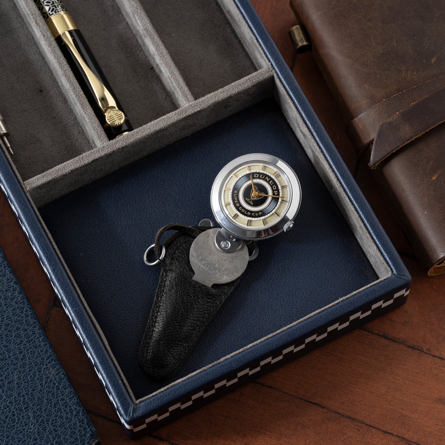 A close-up of the Dunlop Prestige Watch Co. Dunlop Key Watch beside a brown leather journal in a blue and grey case—a perfect gift for any classic car enthusiast.