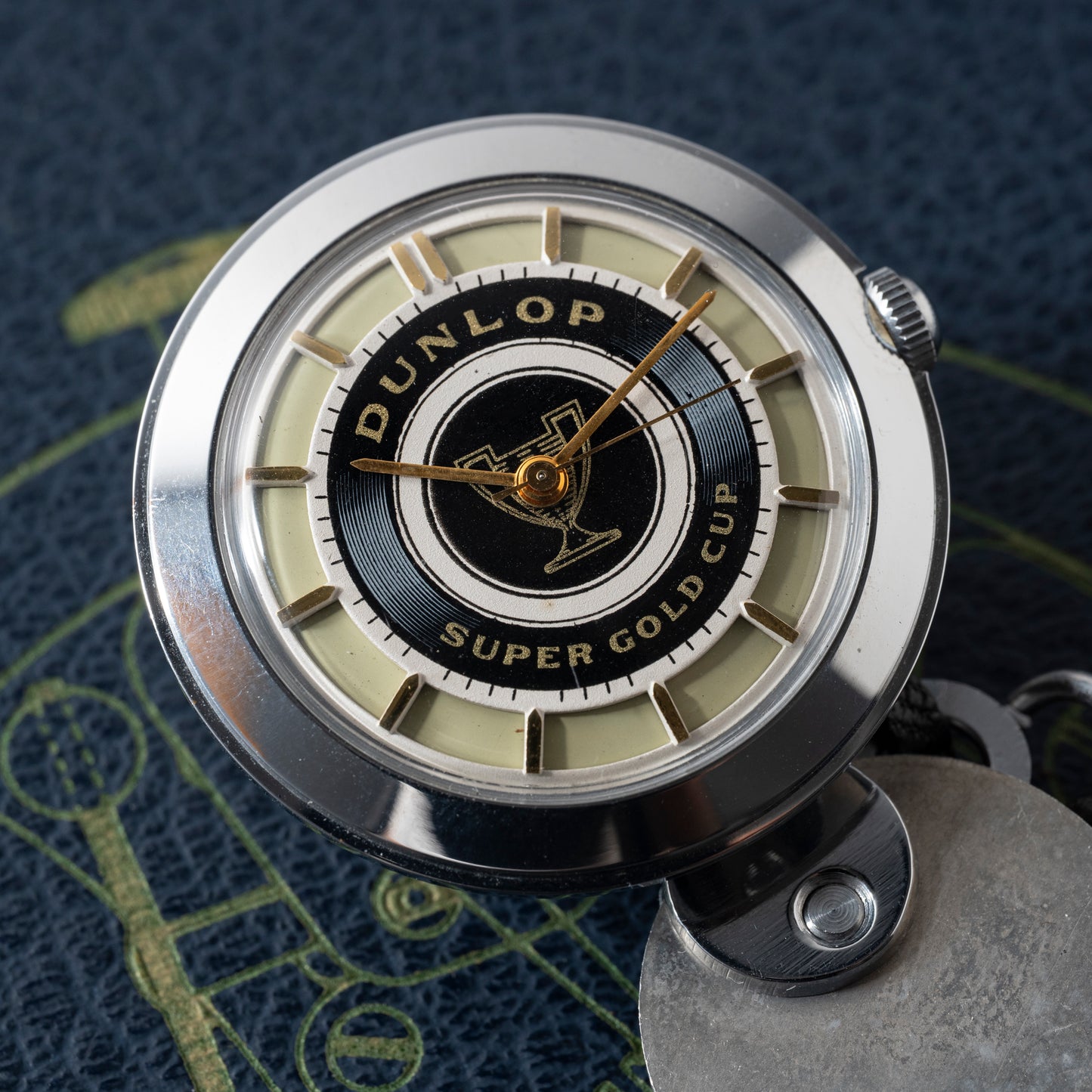 A close-up of the Dunlop Prestige Watch Co. Dunlop Key Watch, featuring a black and gold center inscribed with "Dunlop Super Gold Cup," gold hands, and a silver case, set on a textured dark blue surface with subtle yellow accents.