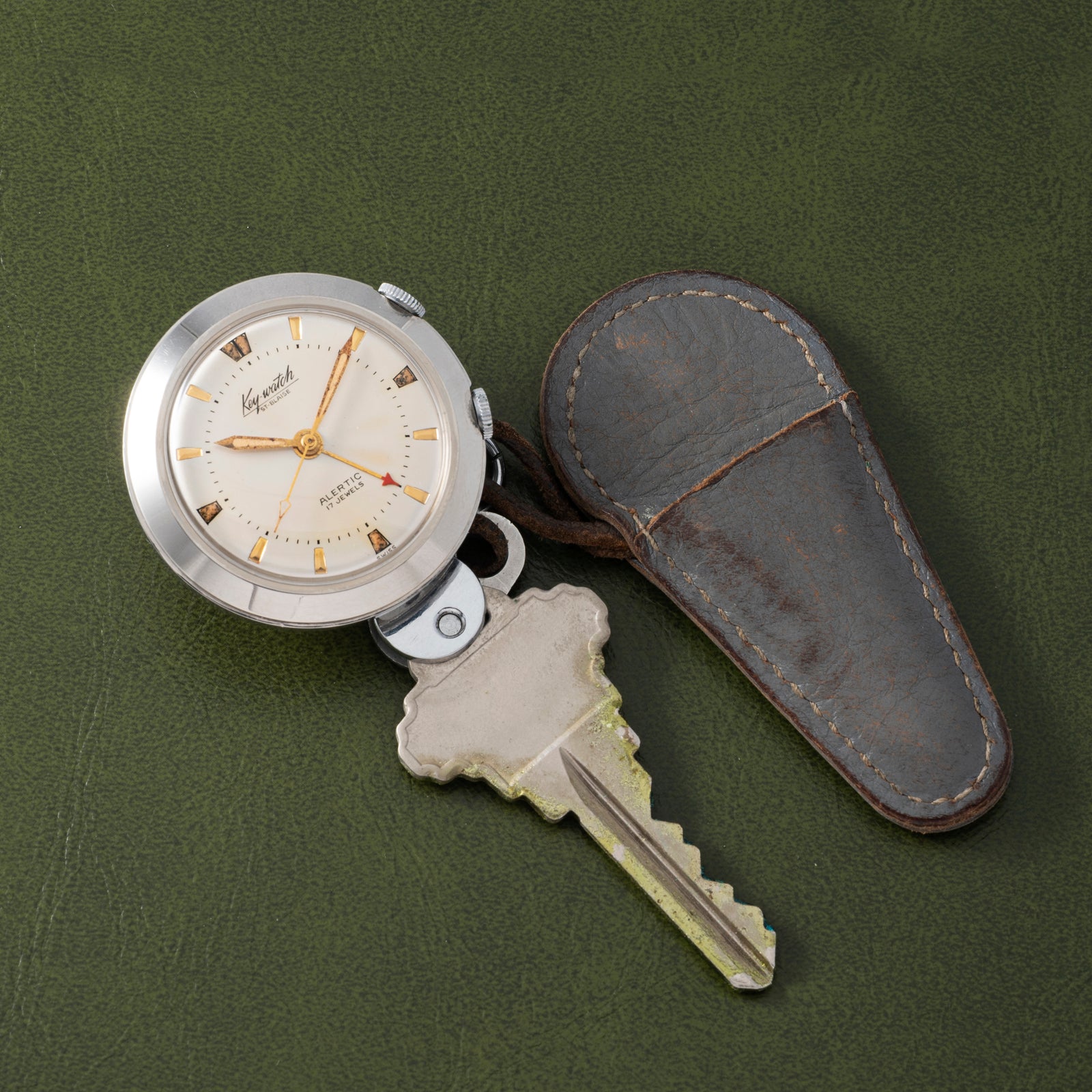 The Saint Blaise Key Watch Alarm, complete with a key and worn leather cover, rests on a textured green surface—an ideal collectible for classic car fans or those who appreciate timeless Saint Blaise treasures.