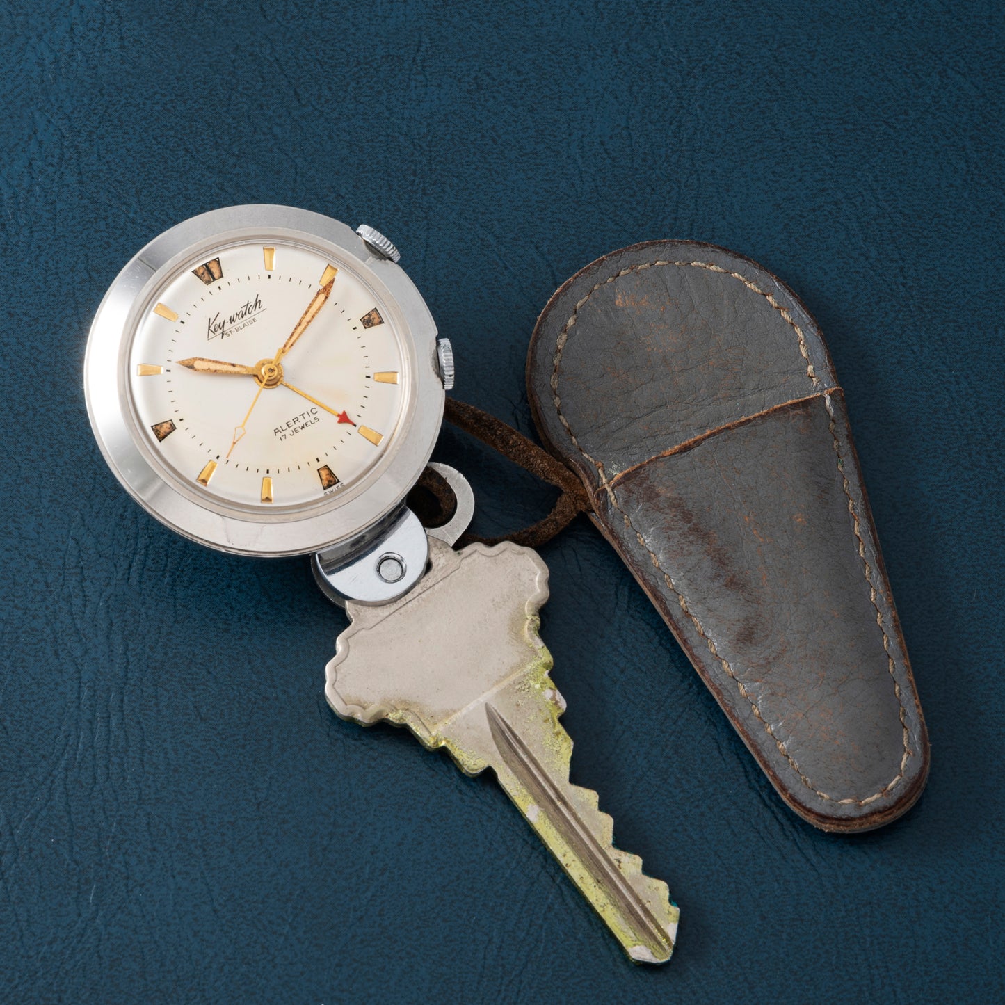 The Saint Blaise Key Watch Alarm by Saint Blaise, featuring Roman numerals and vintage analog design, is attached to a metallic key with a brown leather case, displayed on a dark blue textured surface—ideal for classic car enthusiasts.