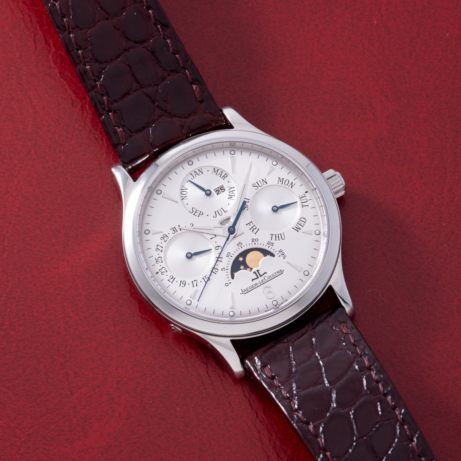 The Jaeger LeCoultre Master Control Perpetual Calendar wristwatch with a brown leather strap, silver case, and white dial featuring subdials and a moon phase display rests on a red textured surface.