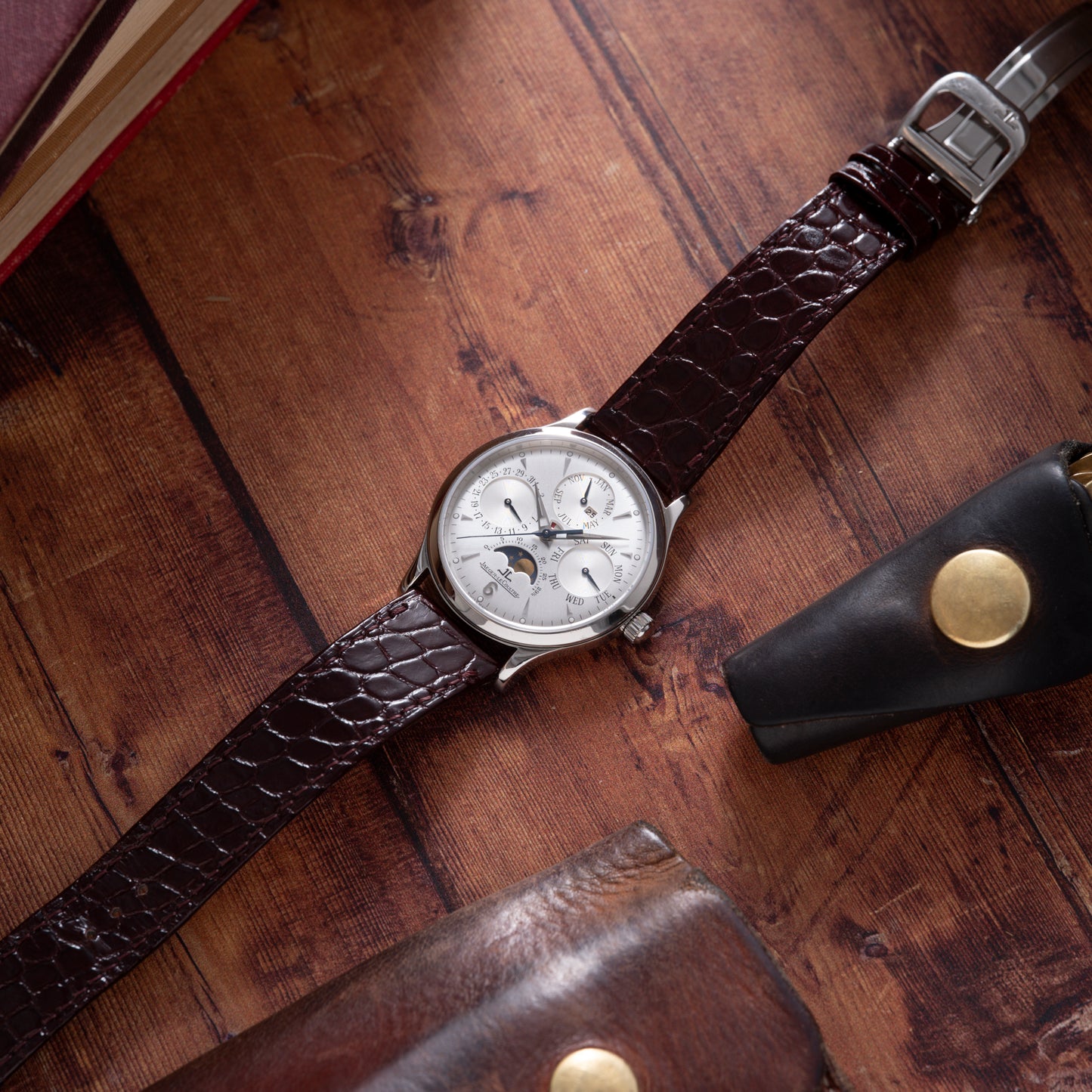 The Jaeger-LeCoultre Master Control Perpetual Calendar, featuring a silver dial with multiple subdials and a brown leather strap, rests on a wooden surface beside a leather case and book.