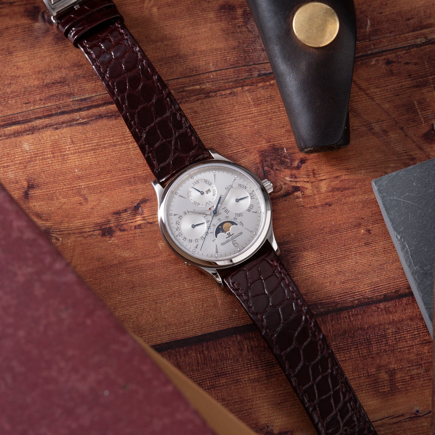 The Jaeger LeCoultre Master Control Perpetual Calendar, featuring a silver case, white dial, and brown leather strap, sits on wood near books and a black leather case with gold button—a timeless classic from Jaeger LeCoultre.