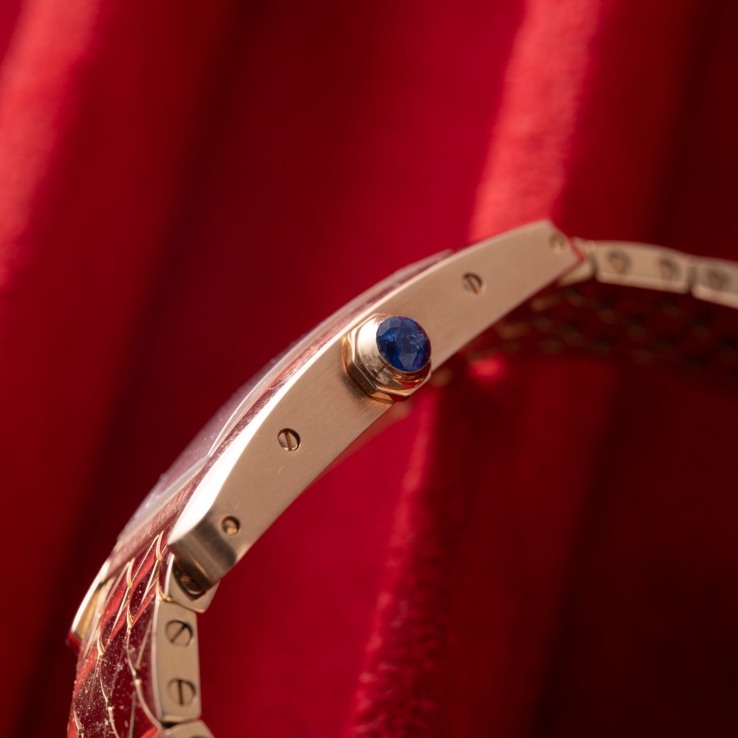 Close-up of a Cartier Tank Americaine ladies 18K rose gold watch by Cartier, featuring a silver guilloché dial and blue gemstone crown, set against red velvet fabric.