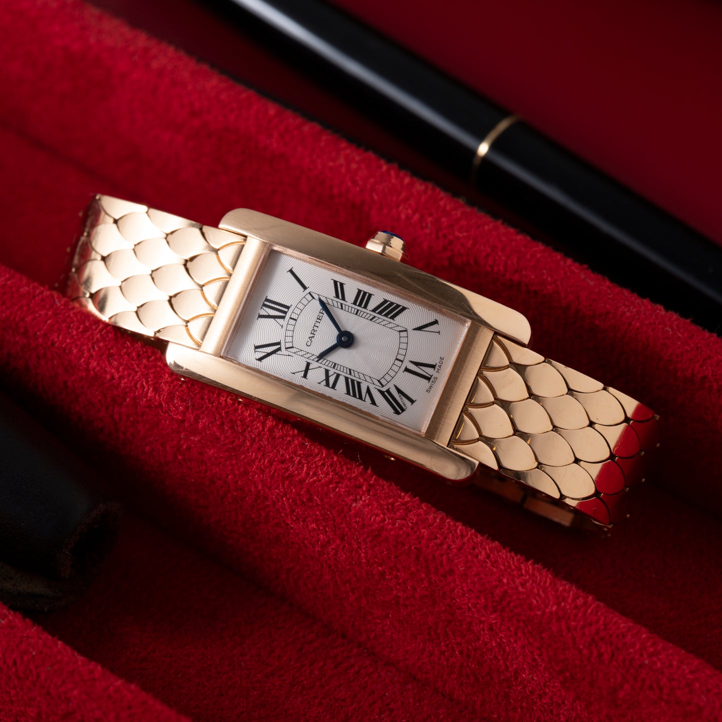 A rectangular ladies' Cartier Tank Americaine in 18K rose gold with a silver guilloché dial and Roman numerals lies on red fabric, with portions of a black pen and leather case visible nearby.