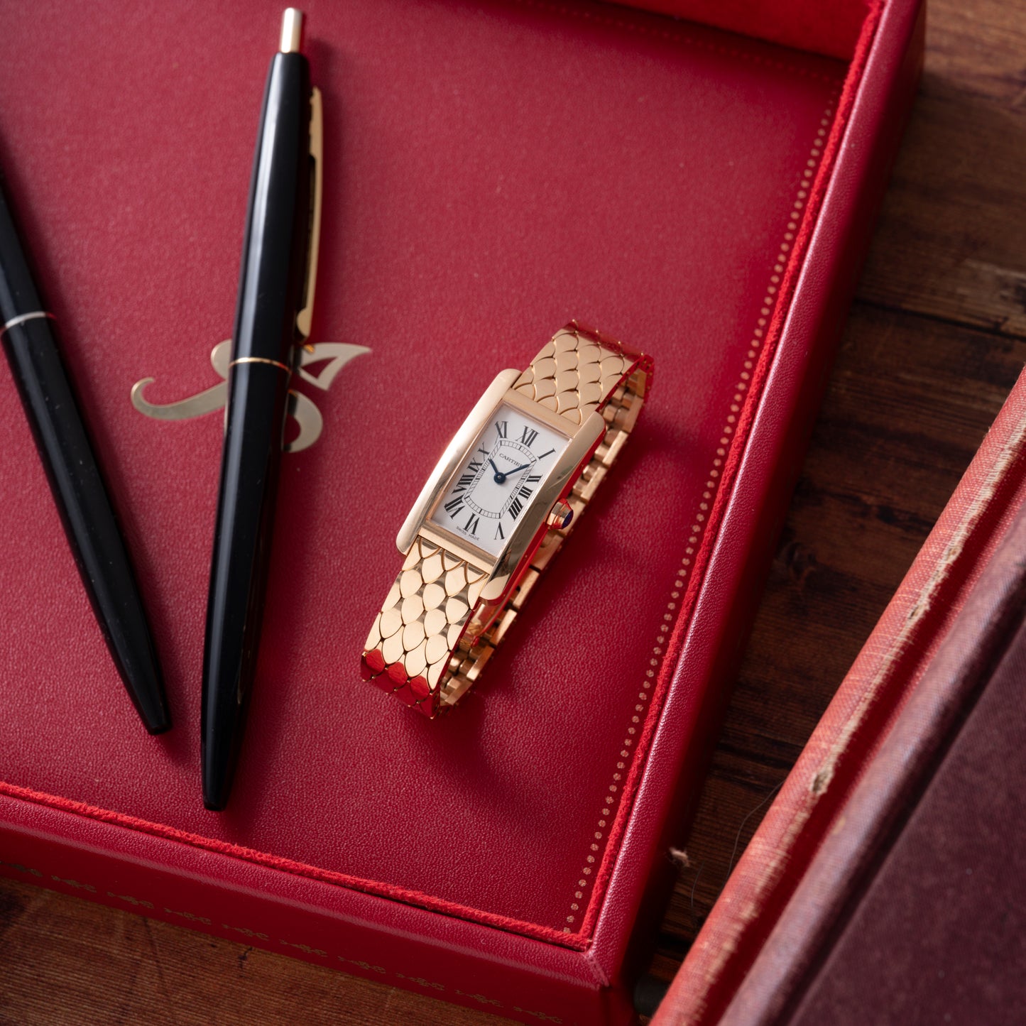 A Cartier Tank Americaine ladies watch by Cartier, featuring an 18K rose gold rectangular face with Roman numerals, rests on a red leather monogrammed box next to two black pens on a wooden surface.