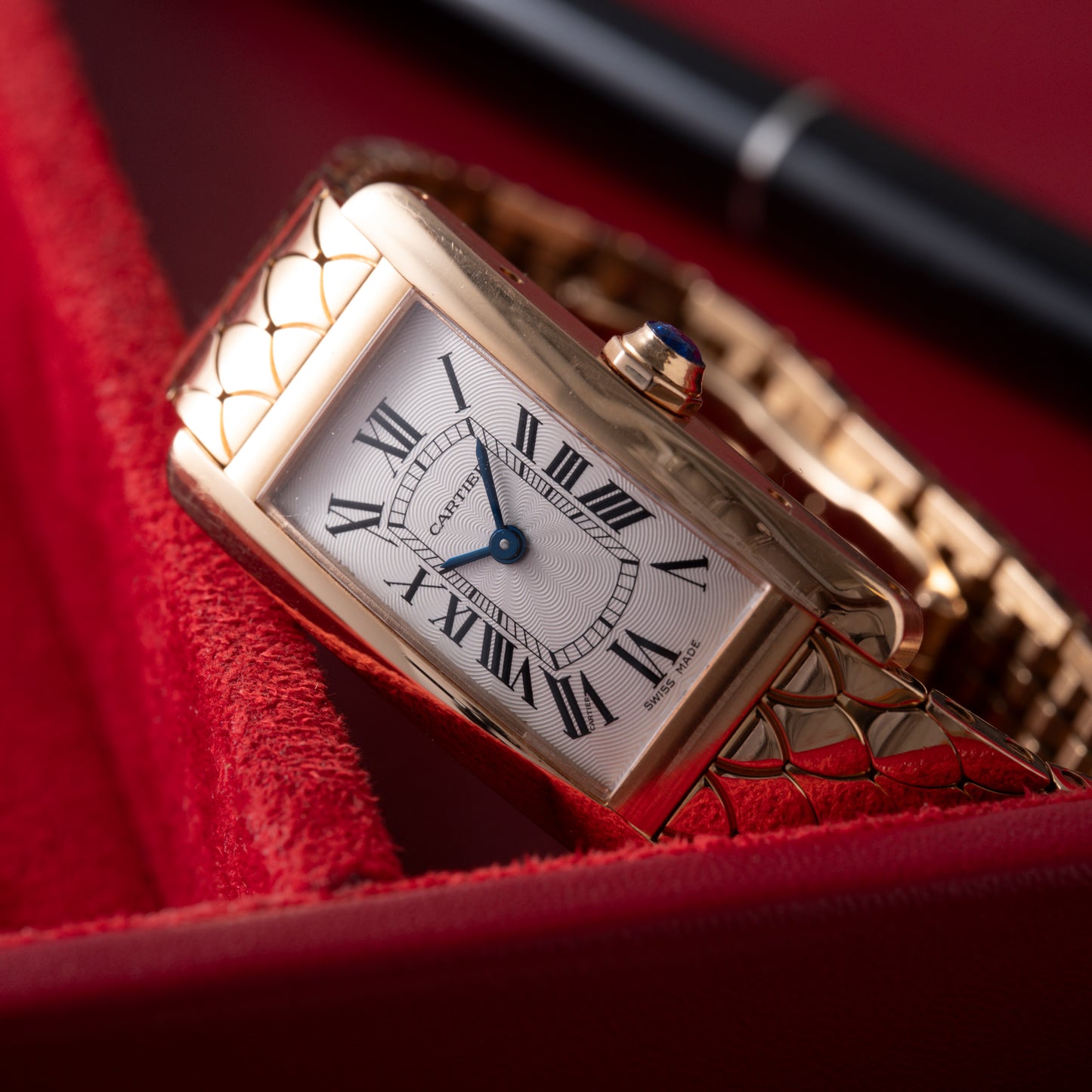 A Cartier Tank Americaine ladies watch by Cartier, with a white face, black Roman numerals, blue hands, and an 18K rose gold scale-pattern bracelet, rests on a red textured surface beside a black pen.