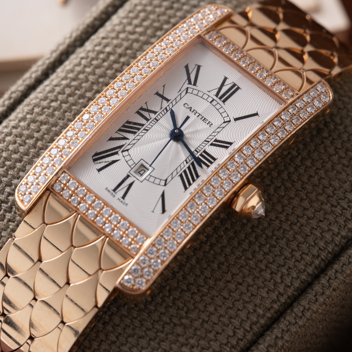 A close-up of the Cartier Tank Americaine Automatic Diamonds watch, featuring a rectangular face, diamond accents, Roman numerals, and a gold bracelet, displayed on a textured fabric cushion.