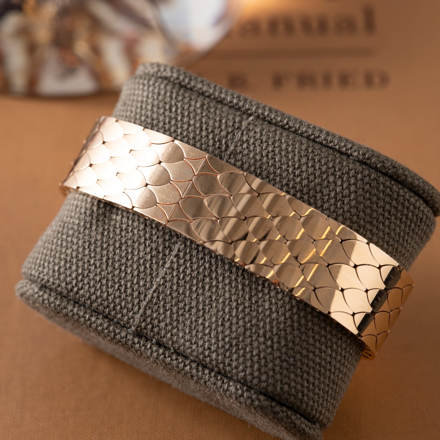 The Cartier Tank Americaine Automatic Diamonds bracelet, featuring a scale-like pattern, is showcased on a textured gray cushion with a blurred brown background.