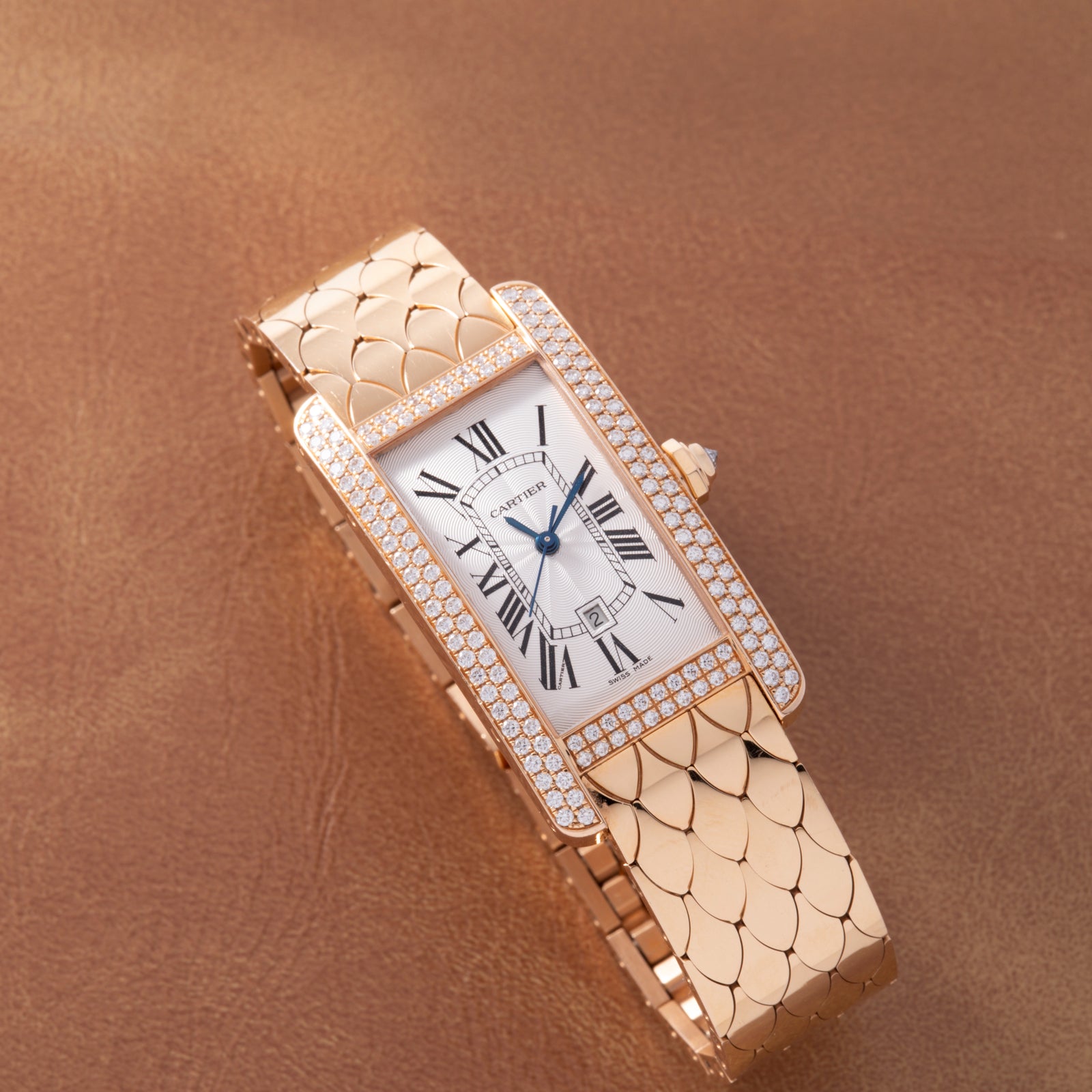 The Cartier Tank Americaine Automatic Diamonds by Cartier features a diamond-set 18K rose gold rectangular case, Roman numerals on a white dial, and a scale-patterned gold bracelet, displayed on brown leather.