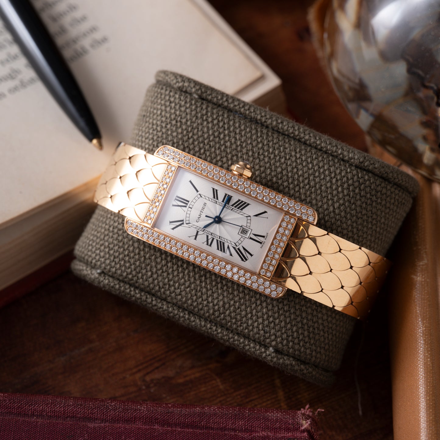 The Cartier Tank Americaine Automatic Diamonds watch, featuring a rectangular face and Roman numerals, is displayed on a textured olive green cushion beside an open book, a pen, and a glass object on a wooden surface.