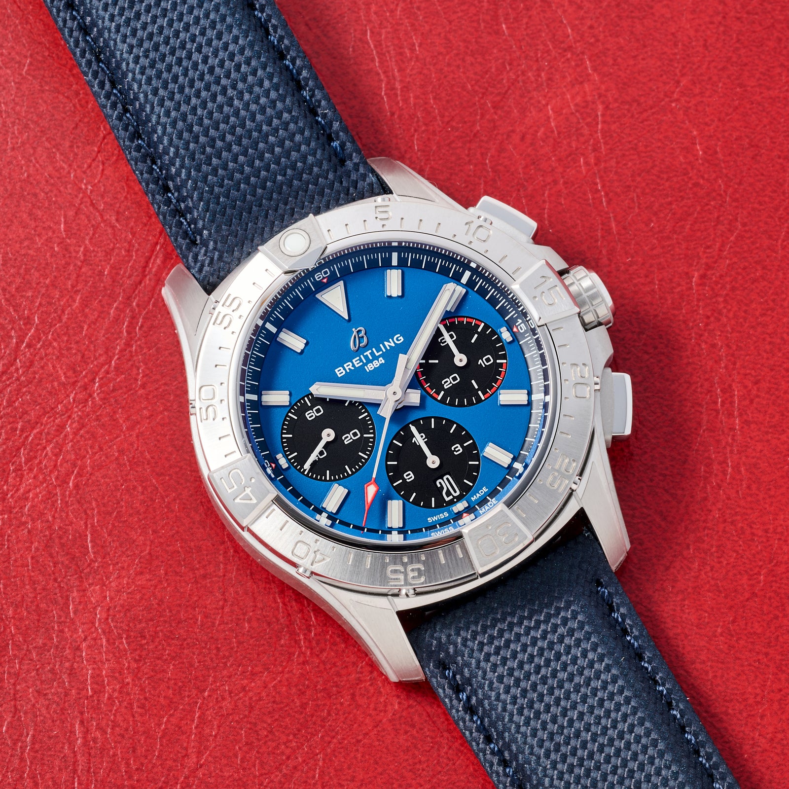 A Breitling Avenger B01 Chronograph 42 with a blue dial, three black subdials, silver indices, and a blue textured leather strap is shown resting on a red leather surface.