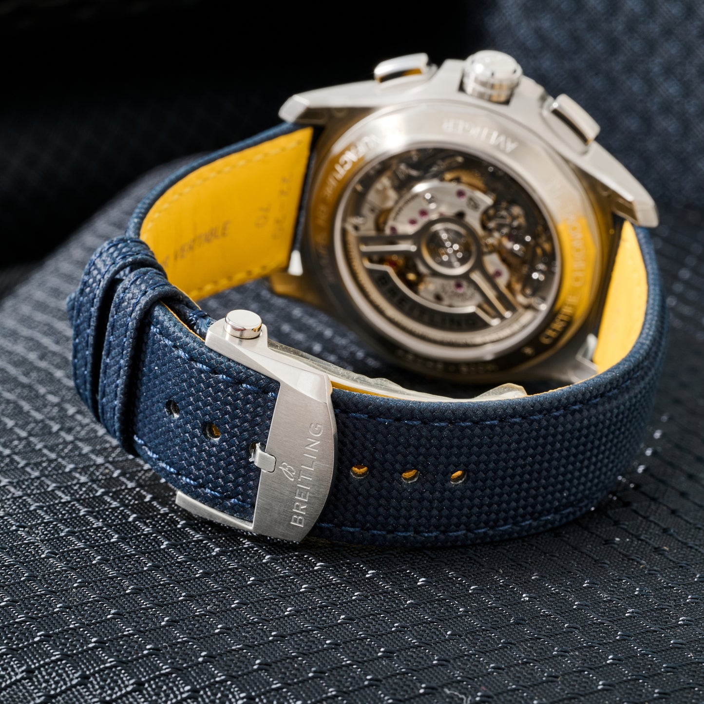 A close-up of the Breitling Avenger B01 Chronograph 42 watch with a blue textured strap and yellow lining highlights its open case back and visible mechanical chronograph movement on a dark, textured surface.