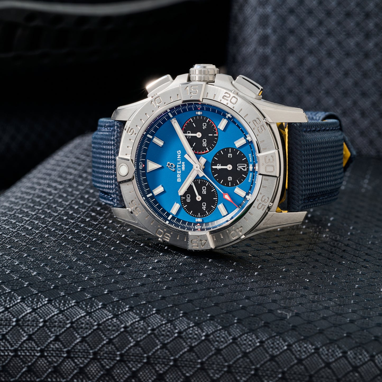 A close-up of the Breitling Avenger B01 Chronograph 42 aviation watch showcases its blue dial, silver bezel, black chronograph subdials, and textured blue strap as it rests on a patterned dark surface.