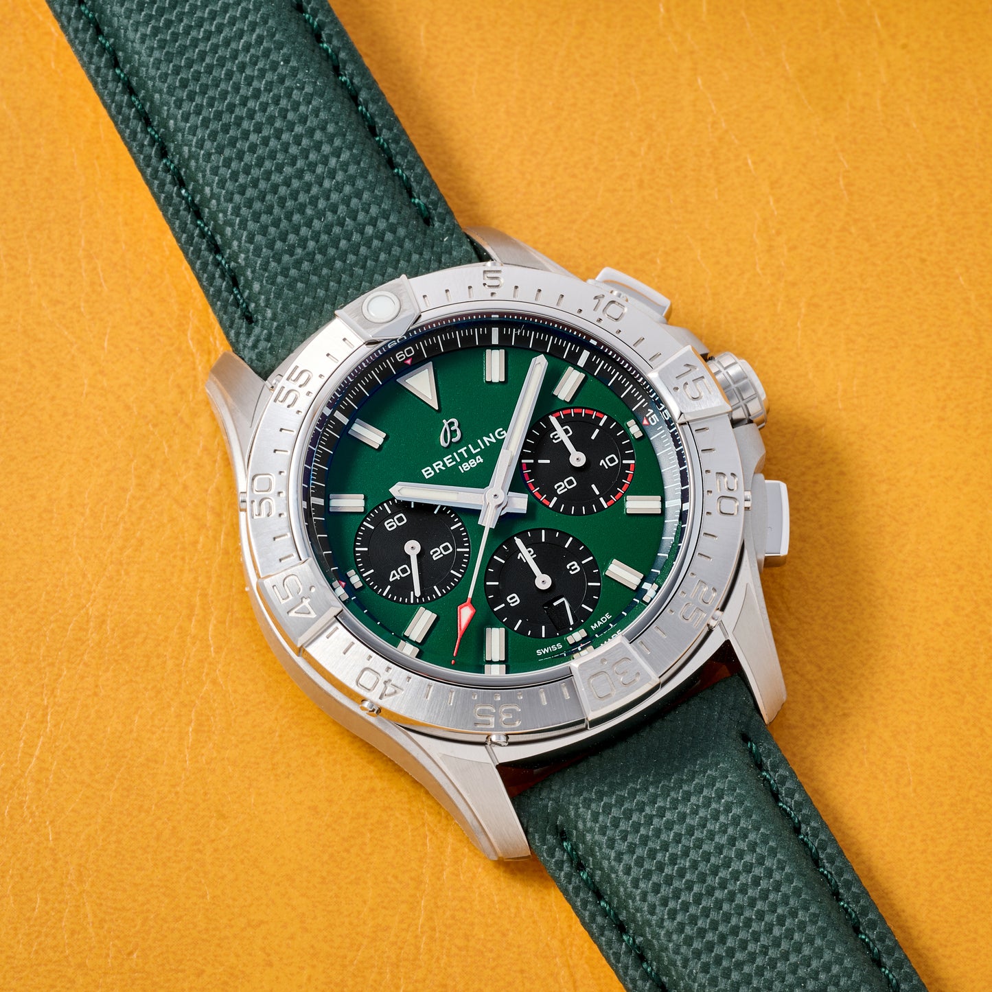A Breitling Avenger B01 Chronograph 42 with a silver case, green dial, and three black subdials rests on a green textured strap atop a yellow leather surface.