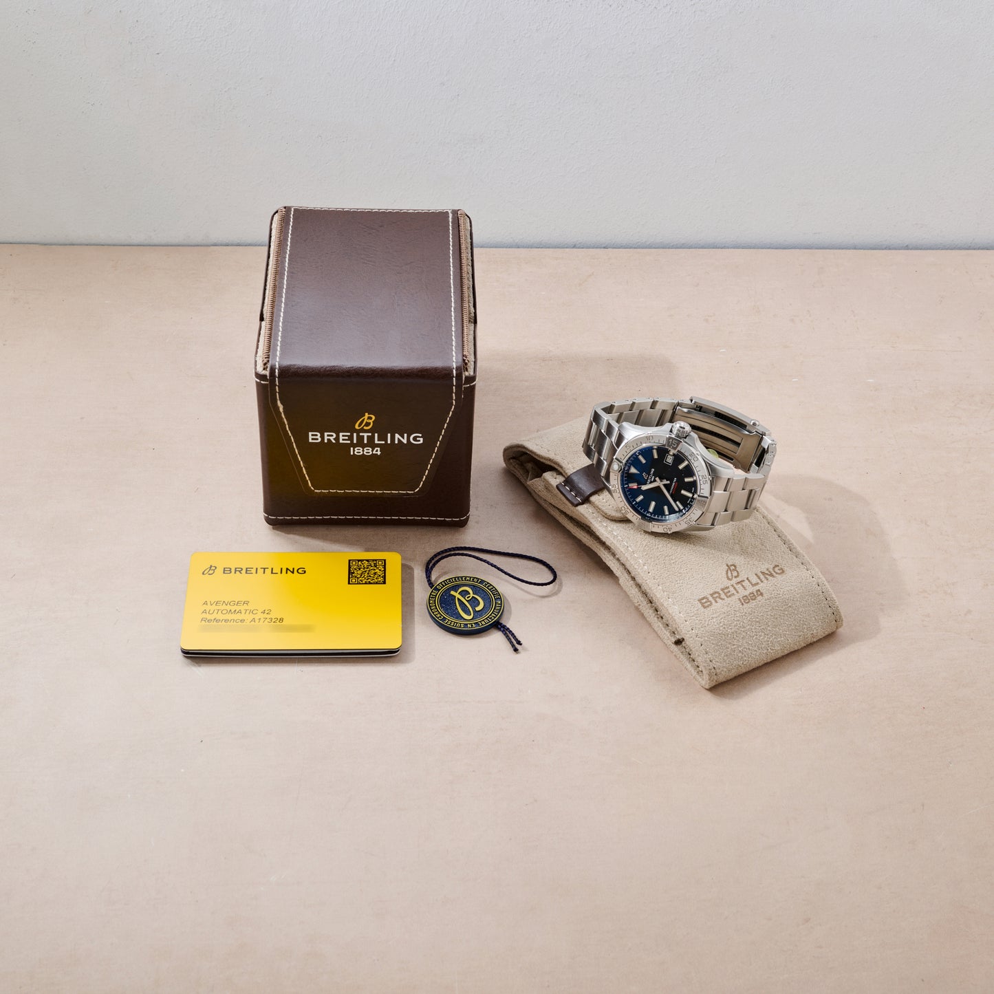 A Breitling Avenger Automatic 42 with a metal band is shown next to its brown box, warranty card, round tag, and beige travel pouch on a light surface.