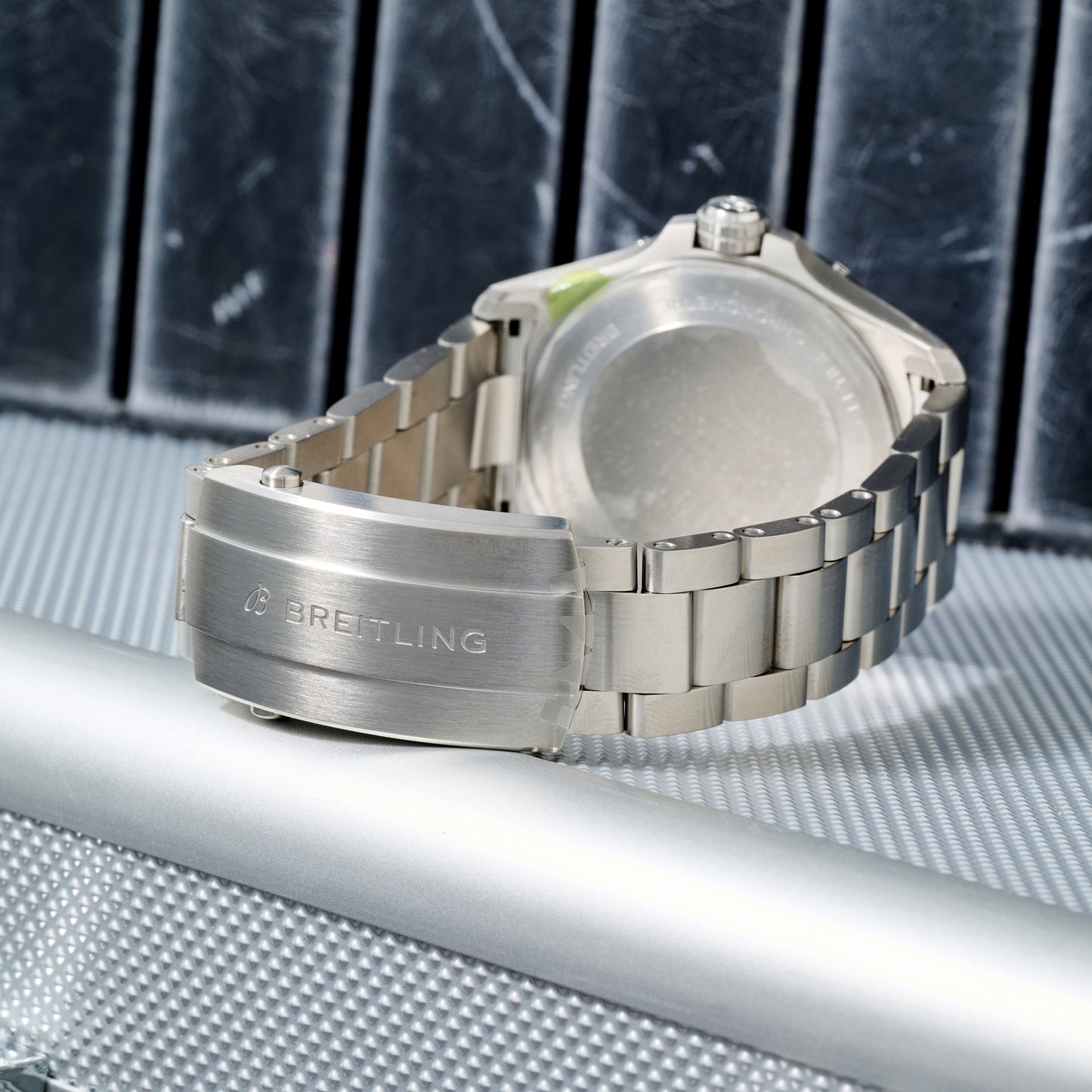 A close-up of the Breitling Avenger Automatic 42 showcases its silver metal bracelet, clasp, and back case on a textured silver surface, with dark panels in the background.