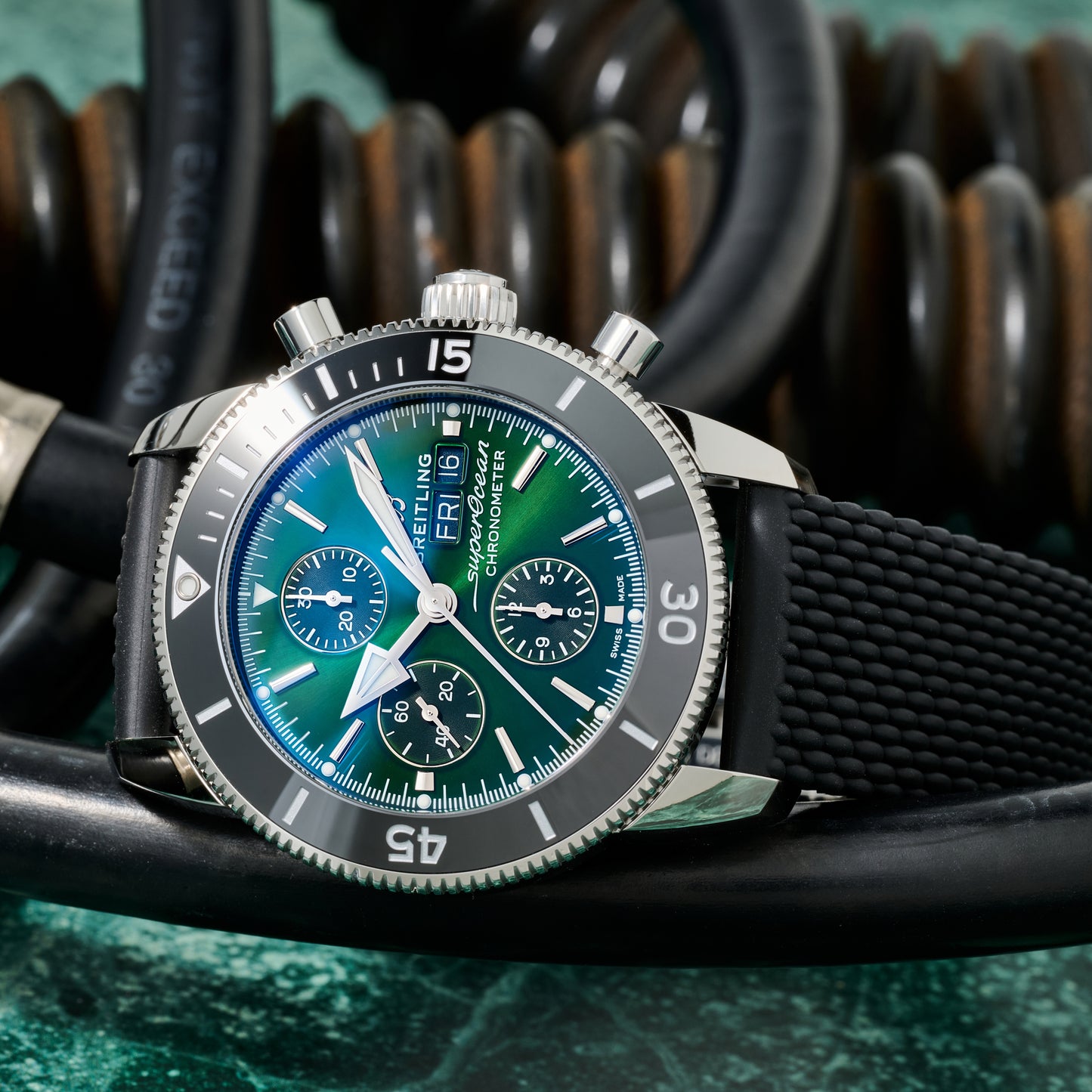 A Breitling SuperOcean Heritage Chronograph 44 with a blue-green dial and black rubber strap rests on coiled black cables, set against a blurred background of springs.