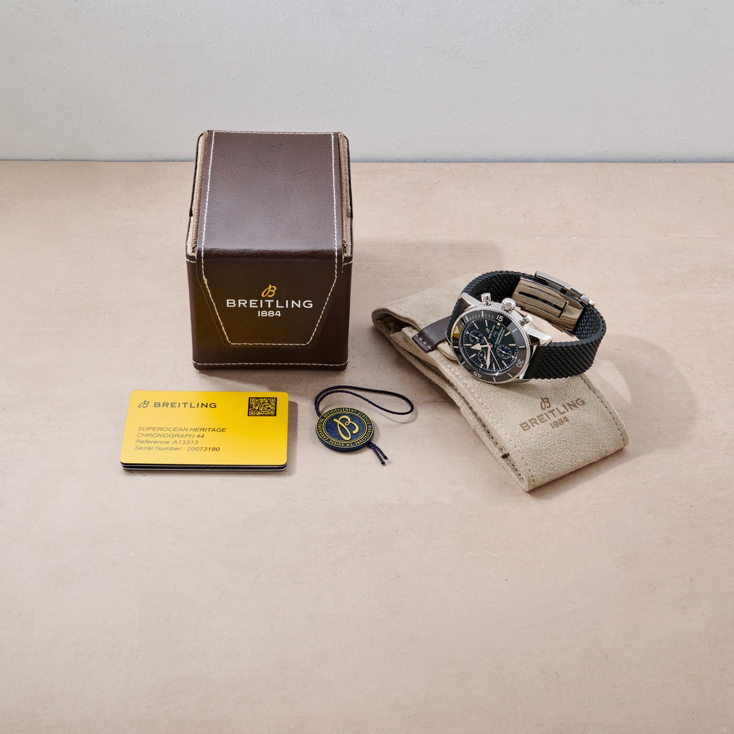 A Breitling SuperOcean Heritage Chronograph 44 by Breitling is shown with its brown box, instruction card, black and yellow tag, and beige travel pouch on a light surface. The watch has a black strap and dark face.
