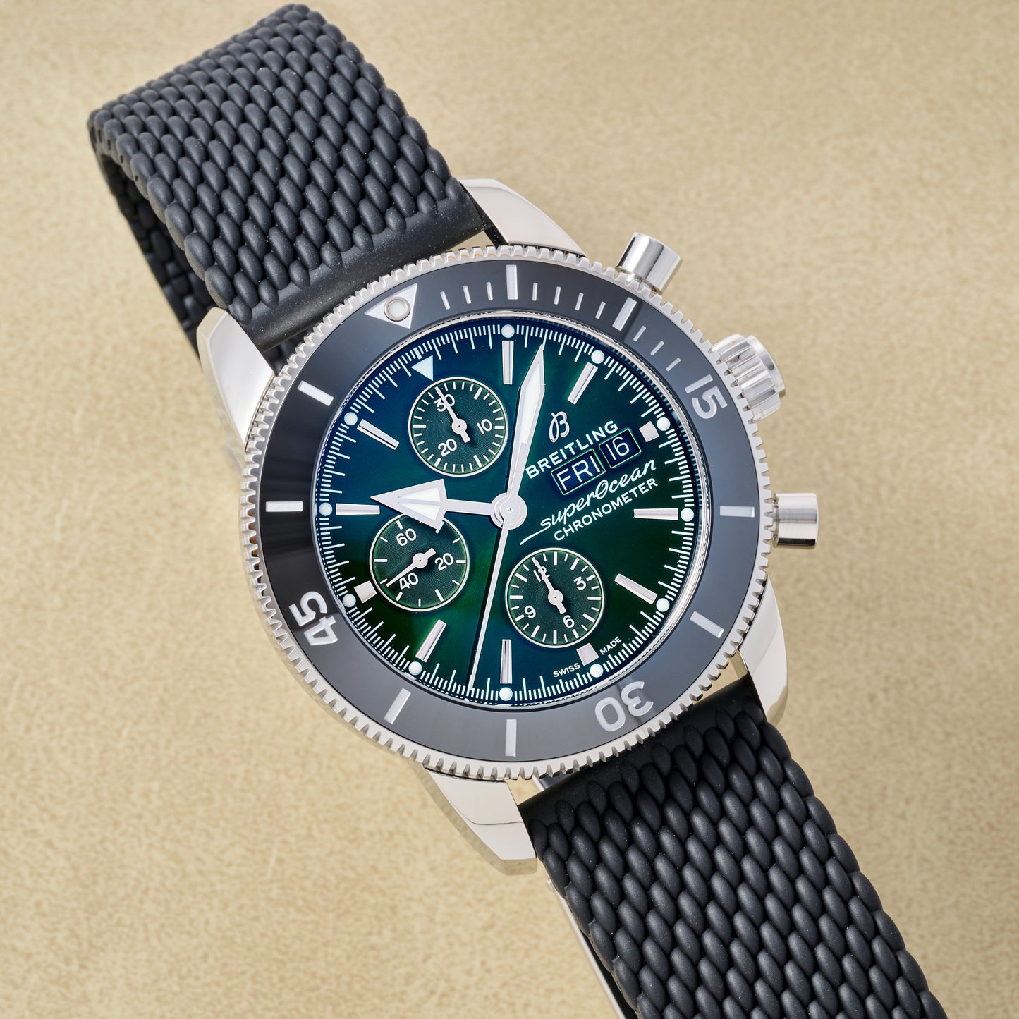 The Breitling SuperOcean Heritage Chronograph 44 features a black textured strap, silver bezel, and three subdials on a dark green dial, elegantly displayed on a beige surface.