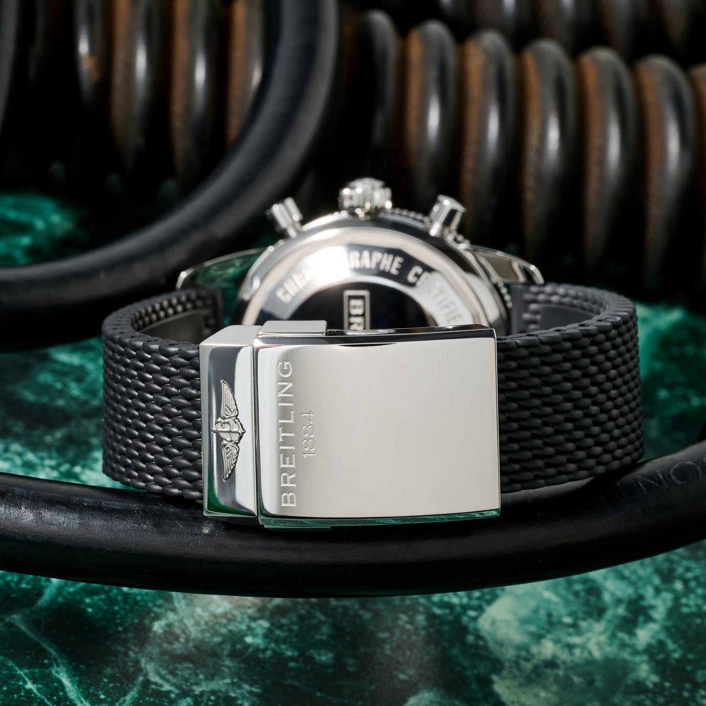 Close-up of a Breitling SuperOcean Heritage Chronograph 44 with a textured black rubber strap and polished silver clasp, set on green marble with coiled black cables in the background.