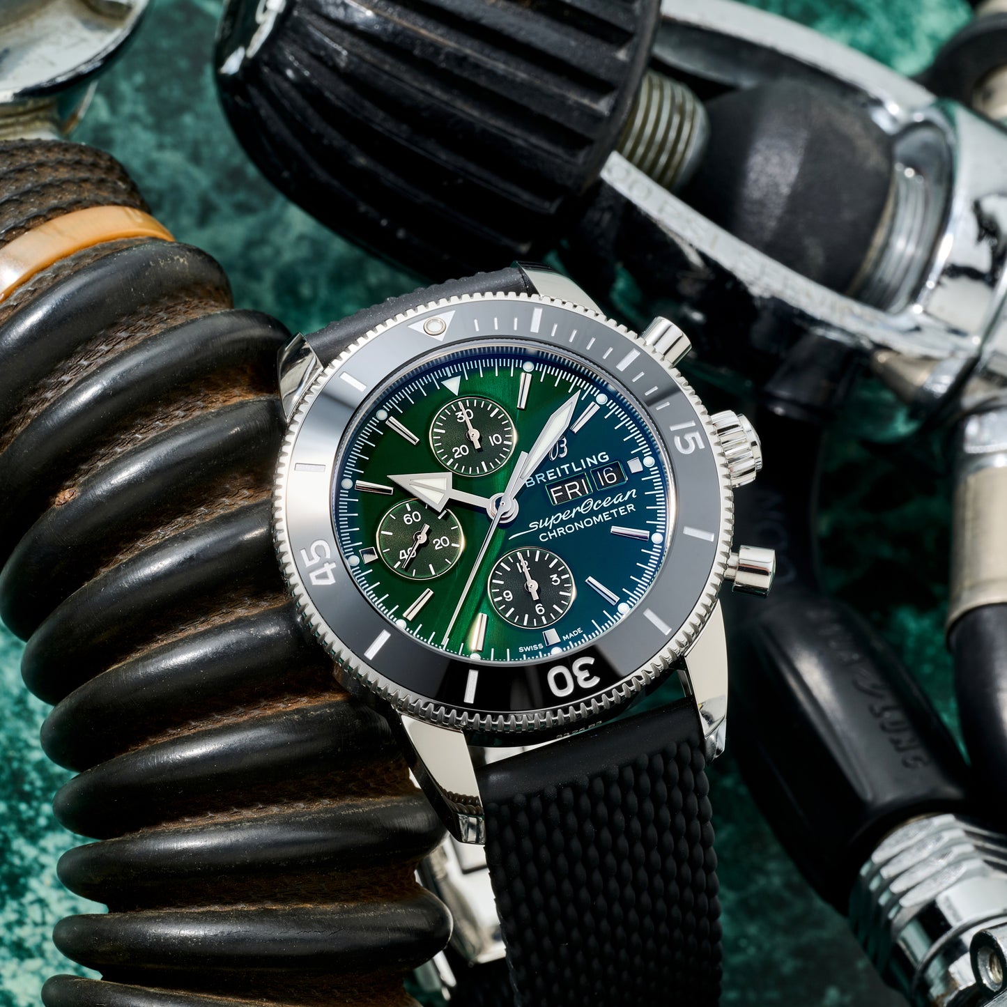 A Breitling SuperOcean Heritage Chronograph 44 with a black rubber strap and blue-green dial is displayed among scuba diving gear, such as breathing hoses and metal regulators.