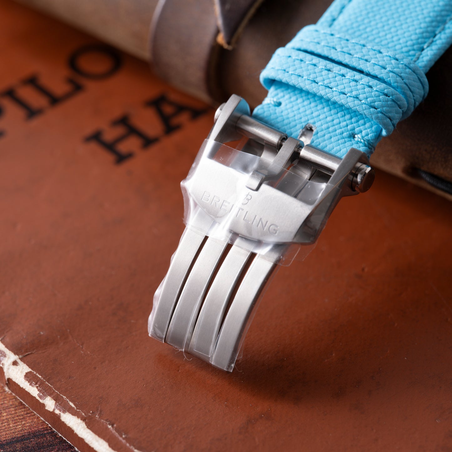 Close-up of a silver Breitling Super Avenger B01 Chronograph 42 clasp on a blue fabric strap, resting atop brown leather-bound books, with "PILOT HAN" partially visible.