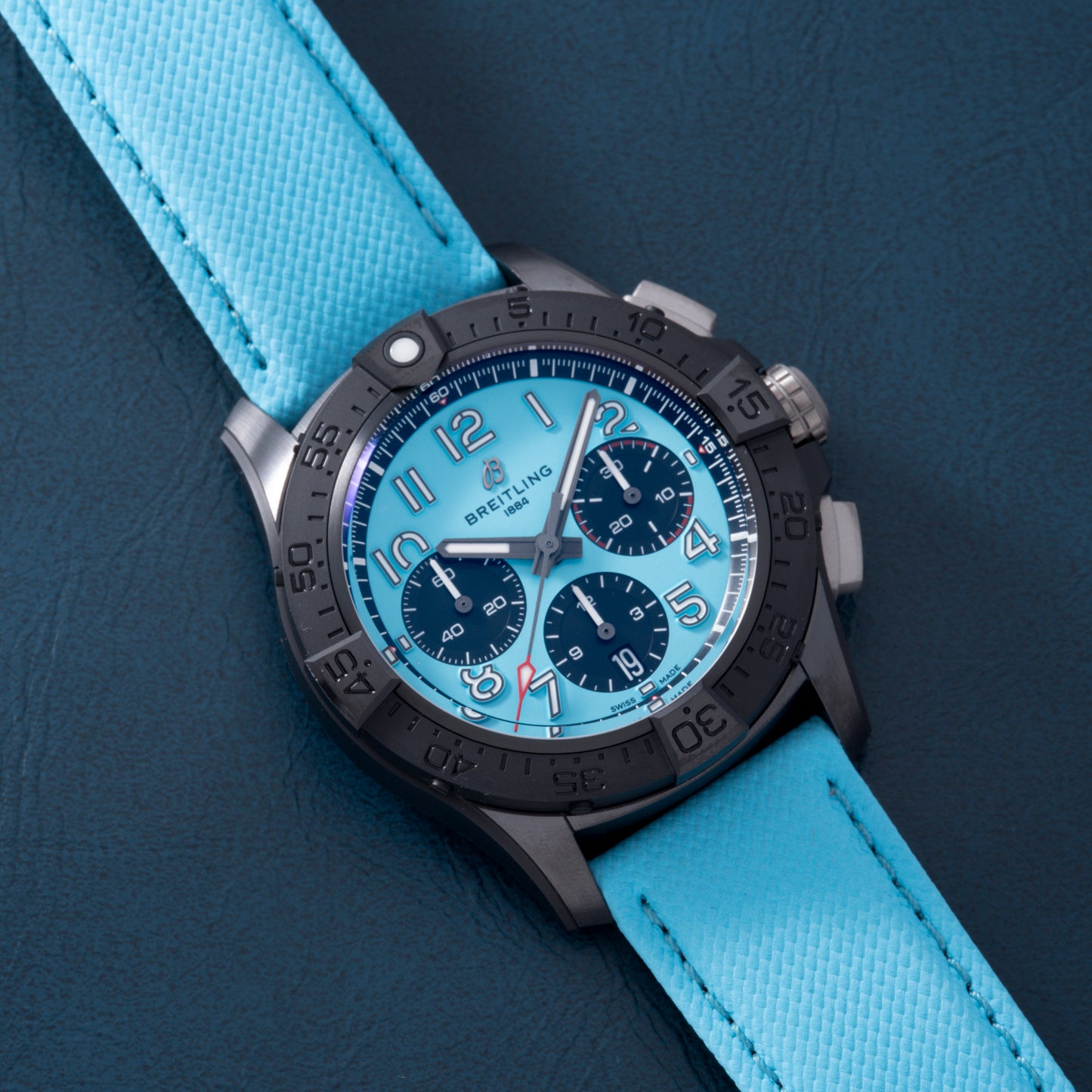 The Breitling Super Avenger B01 Chronograph 42 features a black bezel, light blue dial with three subdials, and a textured strap, elegantly showcased on dark blue leather.