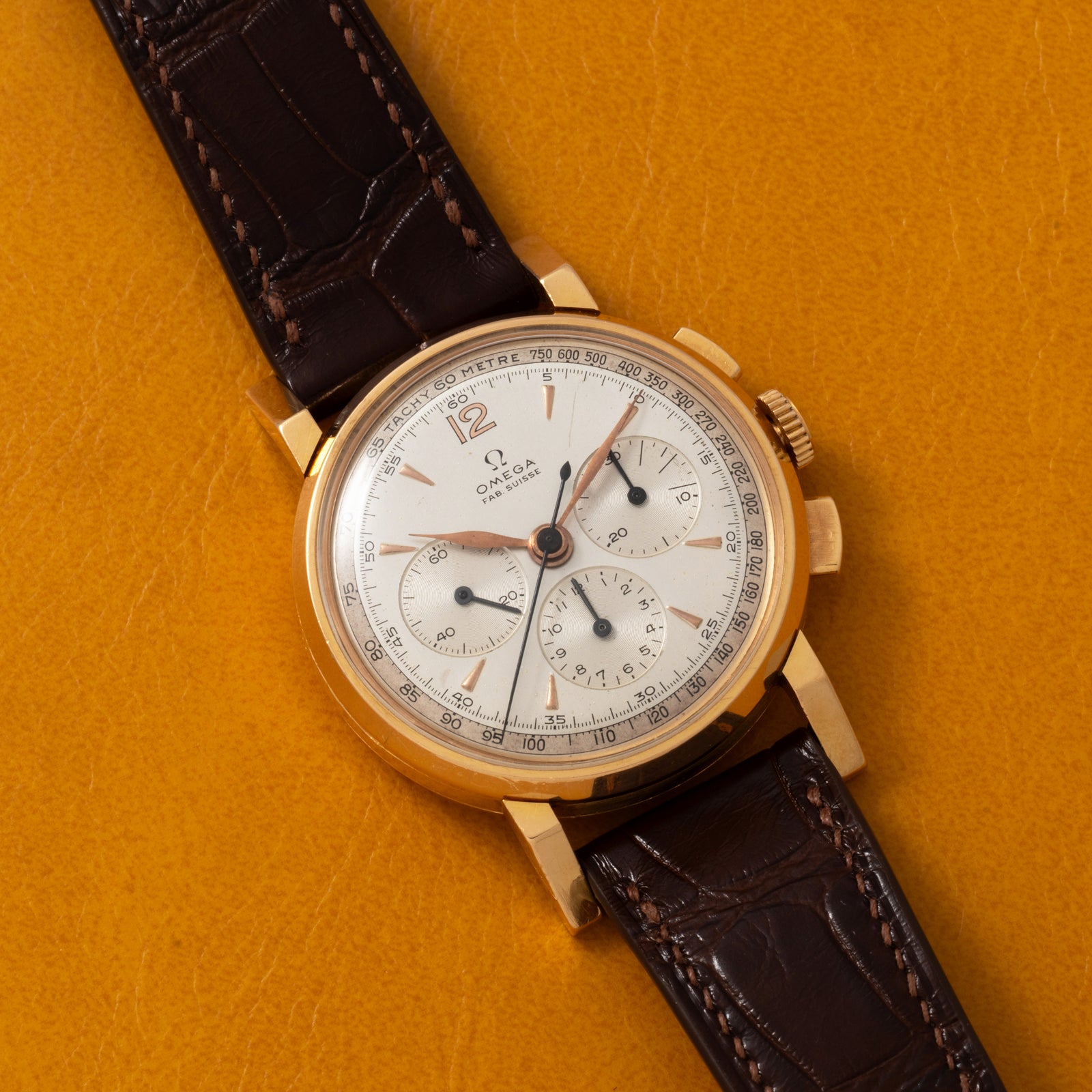 An Omega French Market Chronograph 'Calibre 321' with a gold case, white dial, brown leather strap, three subdials, and gold hour markers rests on a textured mustard yellow surface. It features the renowned Lemania Calibre 321 movement.