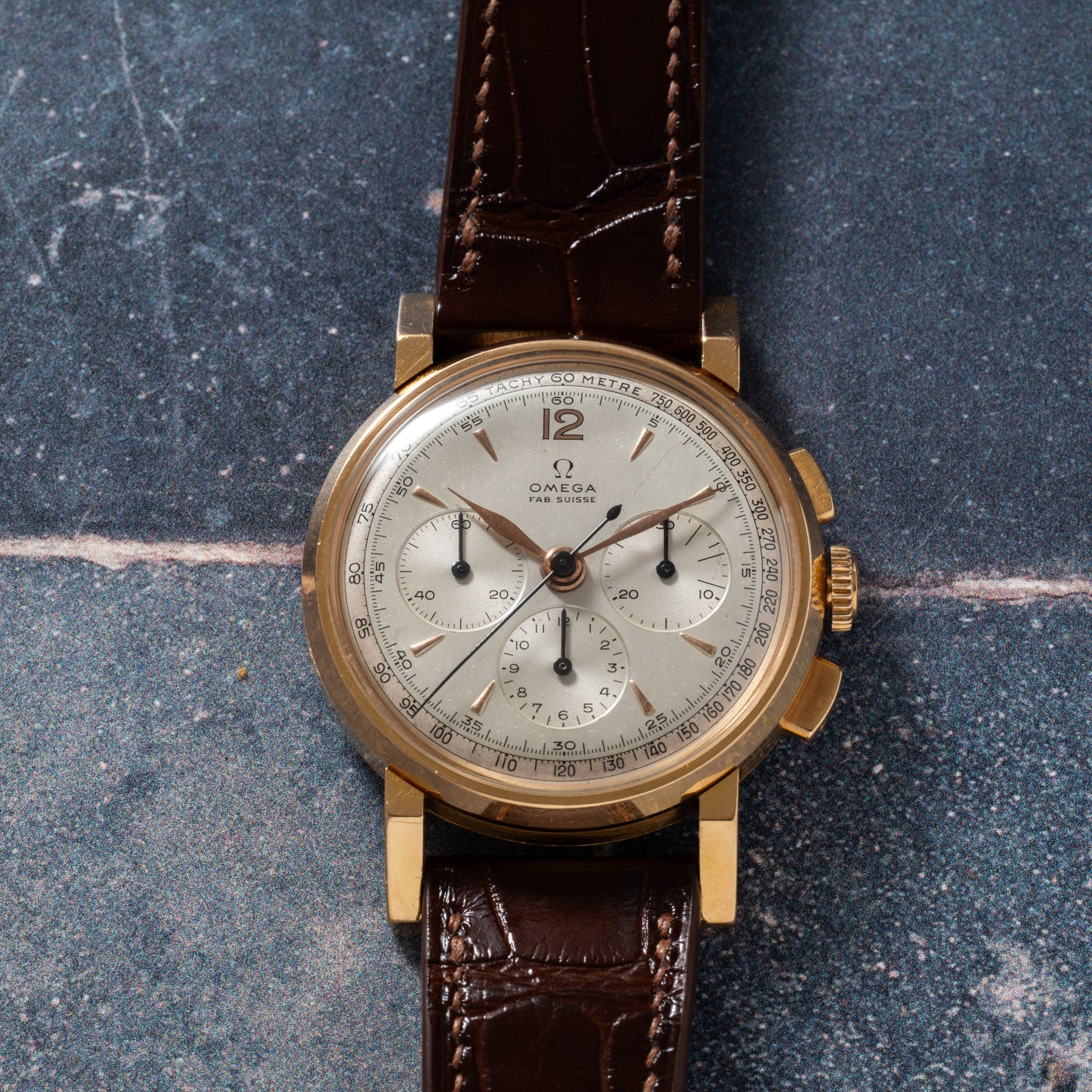 The Omega French Market Chronograph 'Calibre 321' features a gold case, brown leather strap, silver dial with three subdials and black numerals, powered by the Lemania Calibre 321, displayed on a textured blue-gray surface.