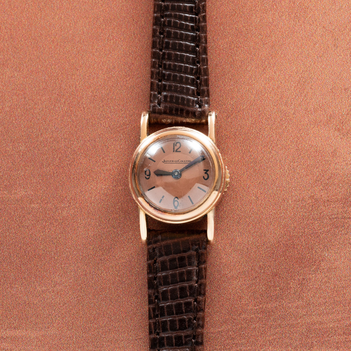 Round rose gold watch with silver dial, Arabic numerals, and dark brown crocodile leather strap on coral background.