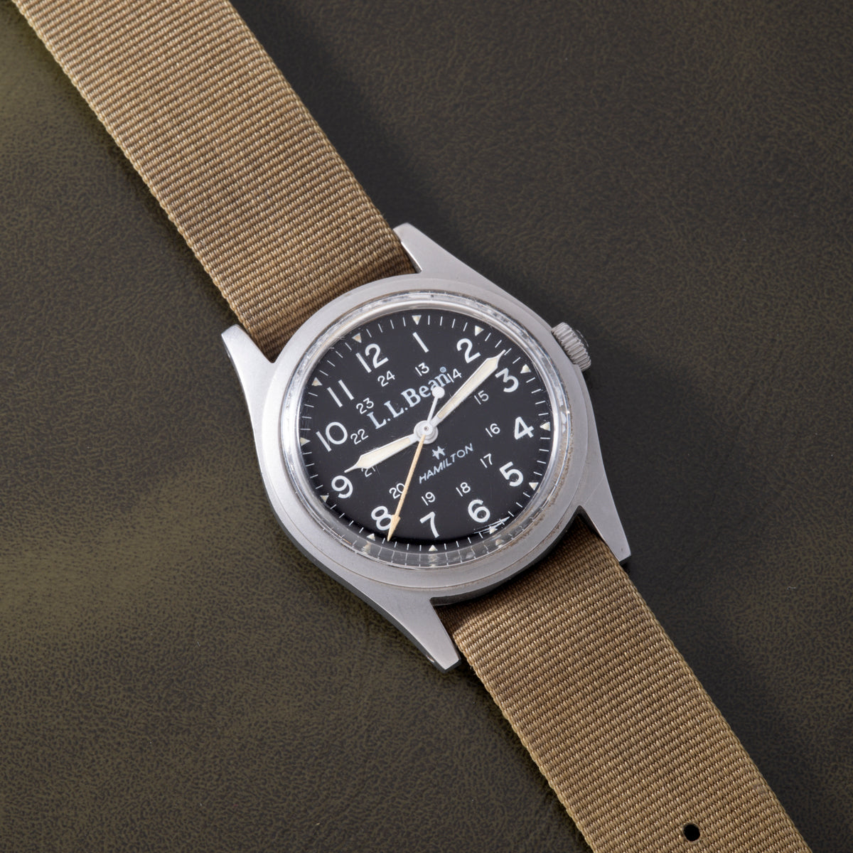 Hamilton Khaki Field mechanical watch with black dial, stainless steel case, and tan fabric strap on neutral background.