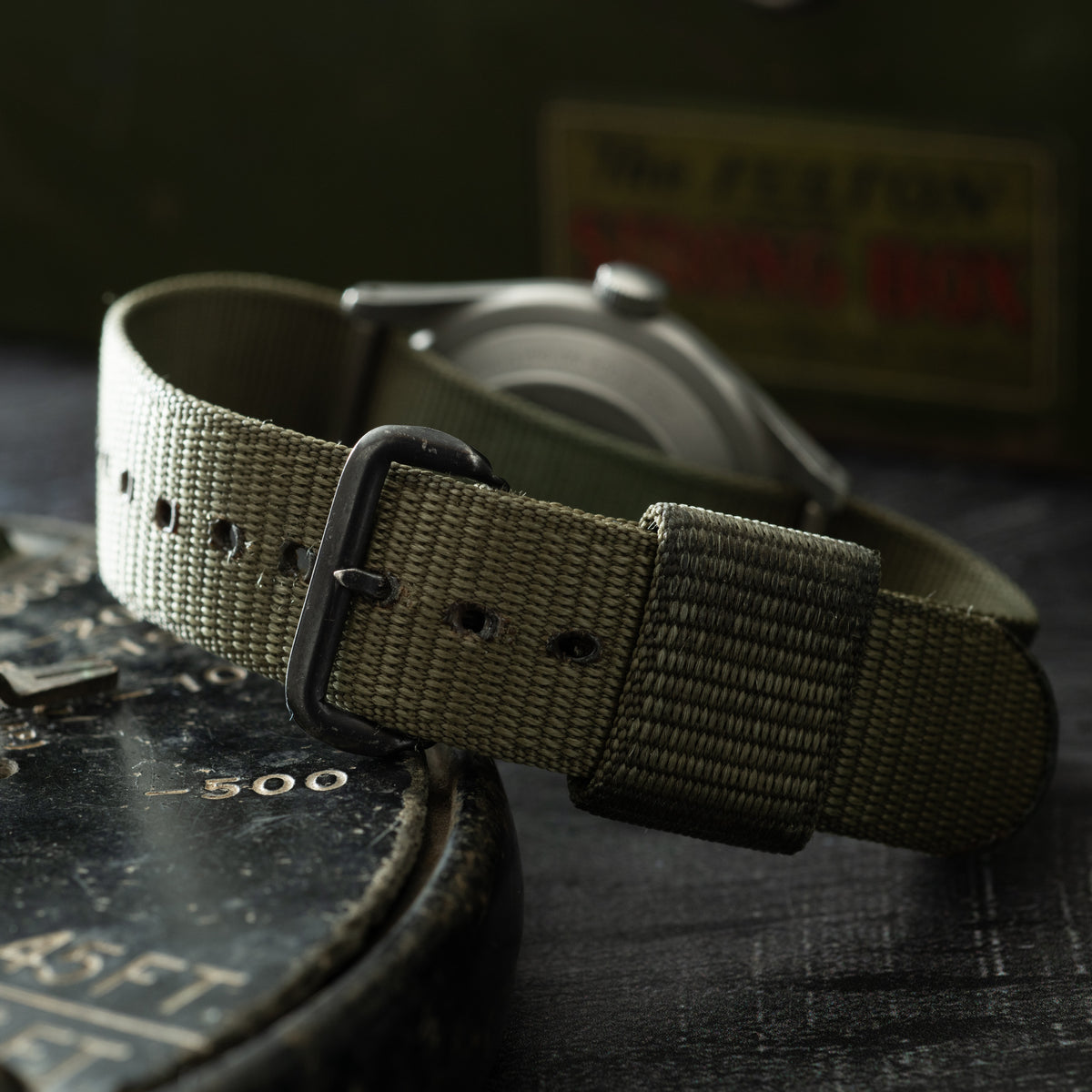 Hamilton GG-W-113 wristwatch with olive canvas strap and black buckle on dark surface.