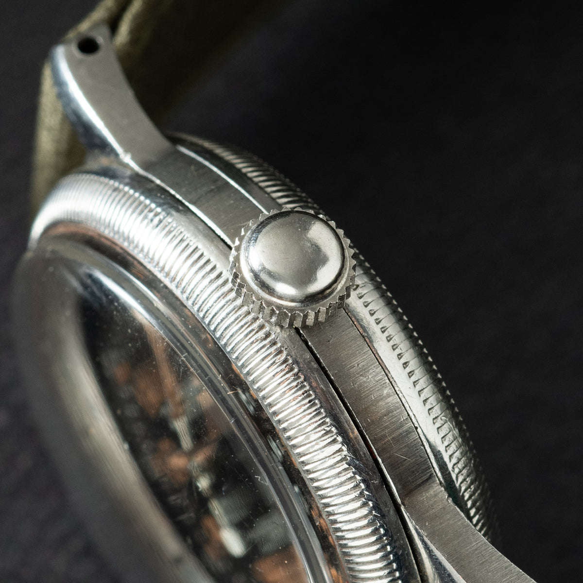 Polished silver-tone watch case with round crown and textured bezel, shown in profile against dark background.