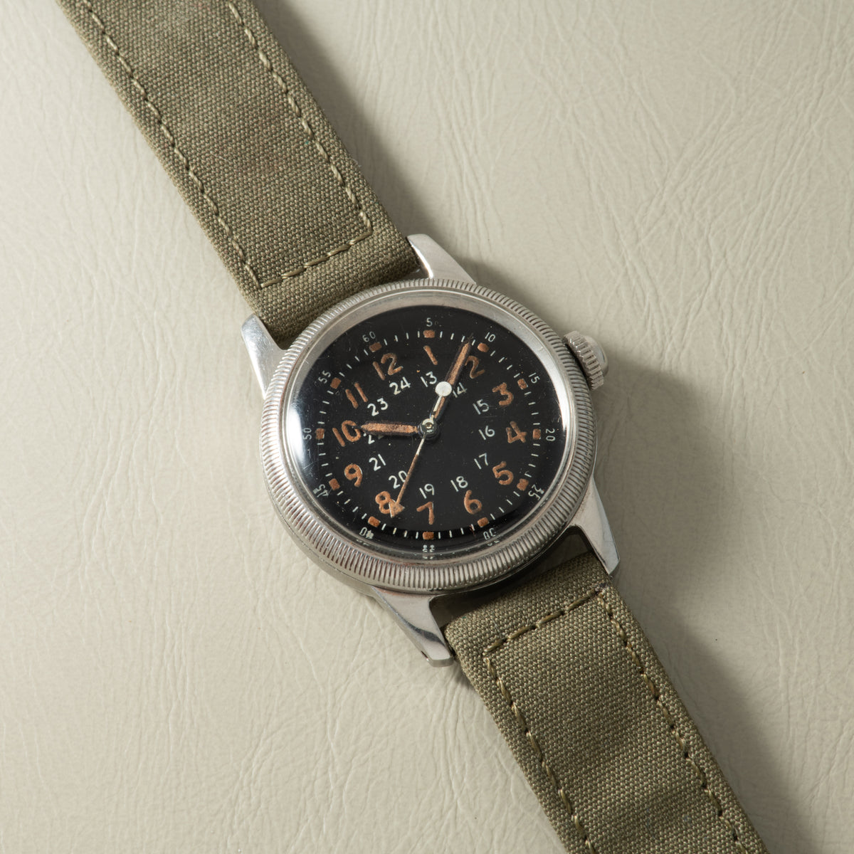 Waltham A-17 watch with black dial, brown numerals, silver case, and tan canvas strap.