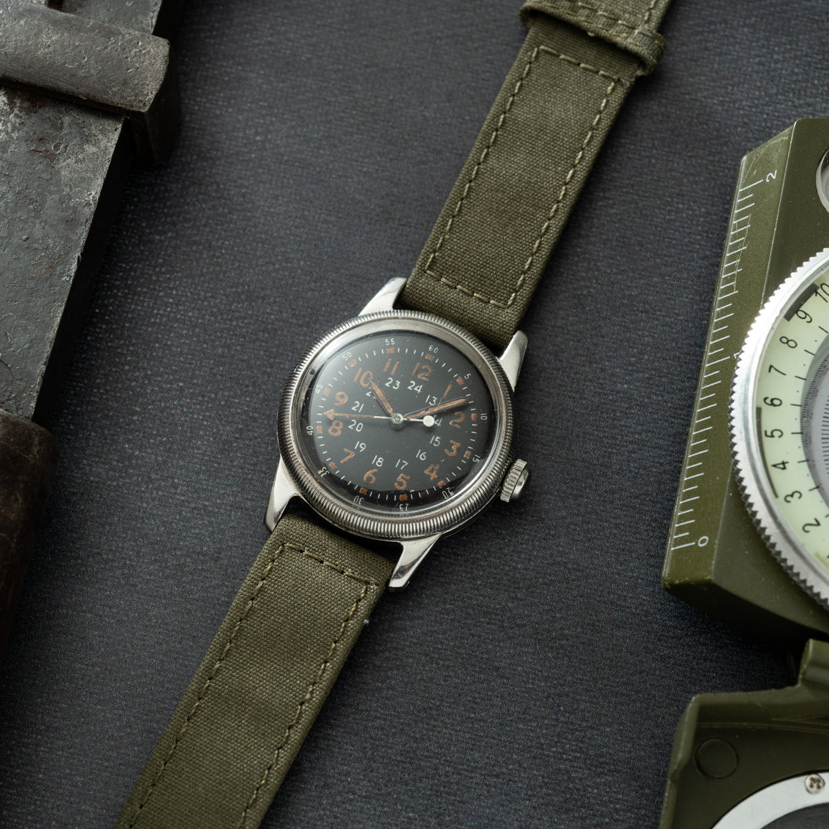 Silver-tone watch with black dial, 24-hour numerals, and olive canvas strap on dark background.