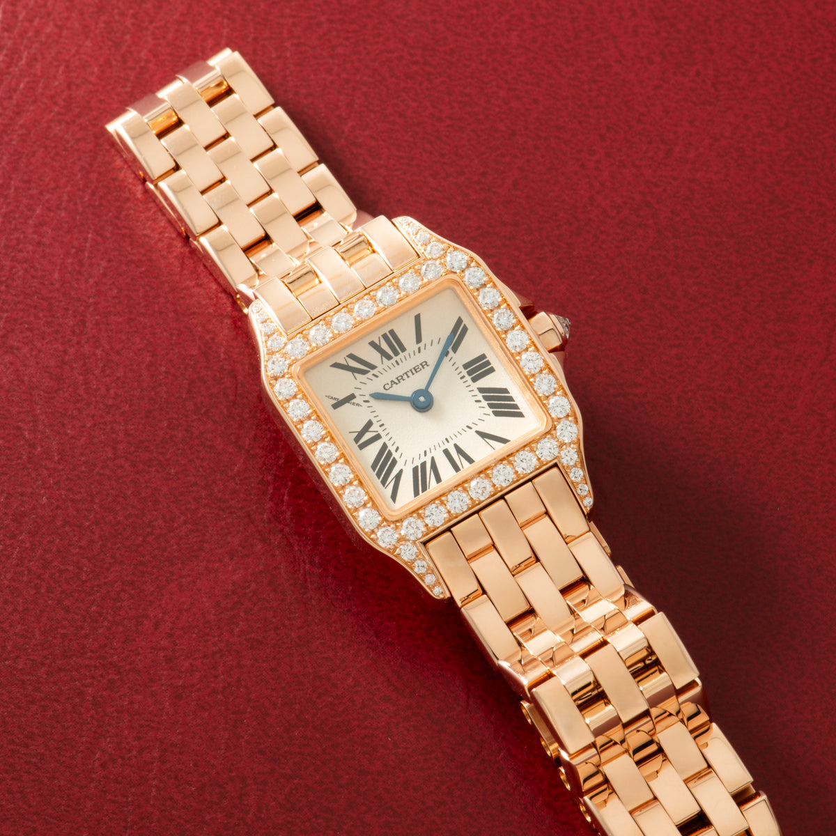 Rose gold square watch with diamond-set bezel, cream dial, Roman numerals, and linked bracelet on red background.