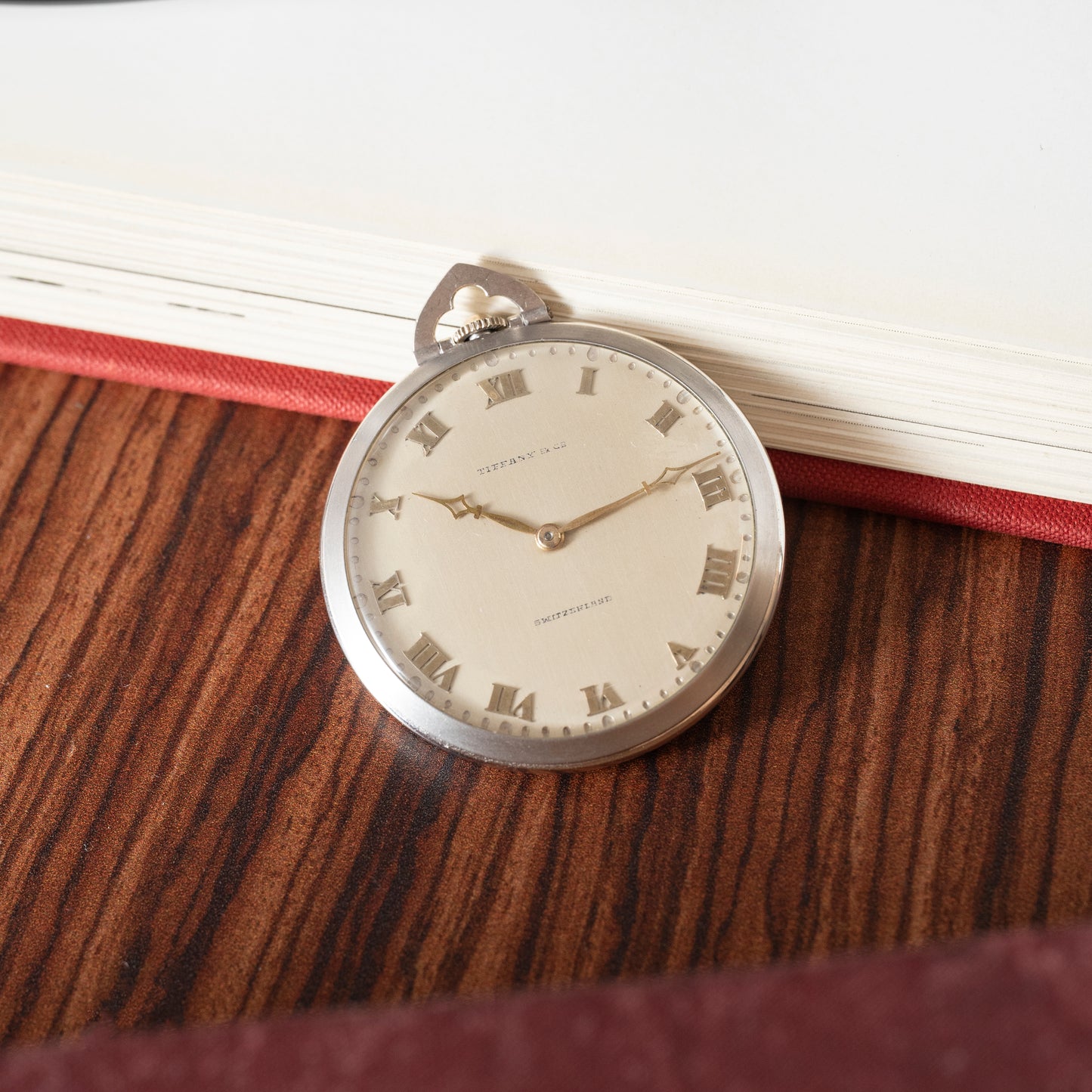 A Tiffany & Co. Platinum Pocket Watch featuring Roman numerals is displayed on a wooden surface beside an open, cream-paged book with a red cover.