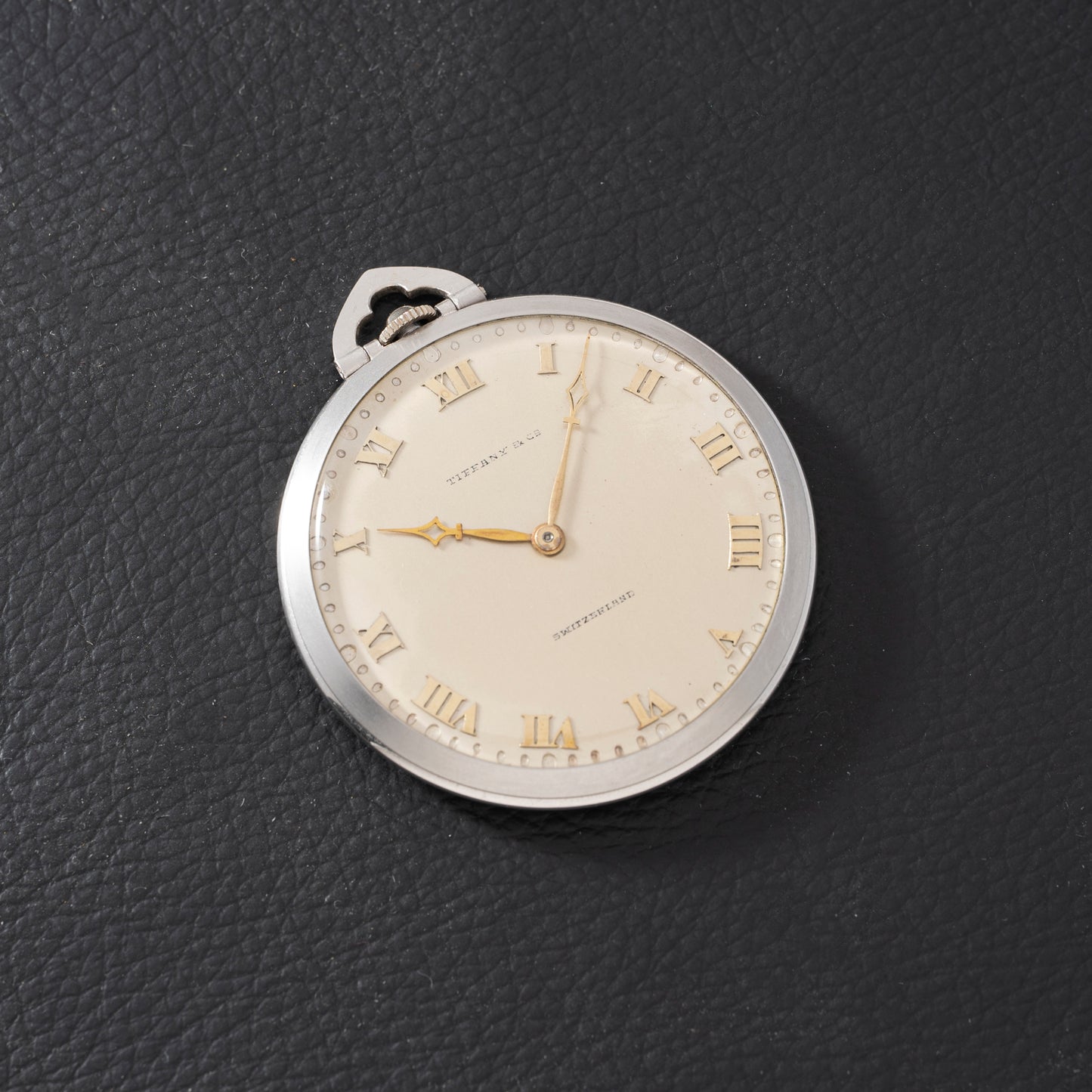 The Tiffany & Co. Platinum Pocket Watch, featuring gold Roman numerals and hands, rests on a textured black surface at 6:17, showcasing the brand's classic mechanical craftsmanship.