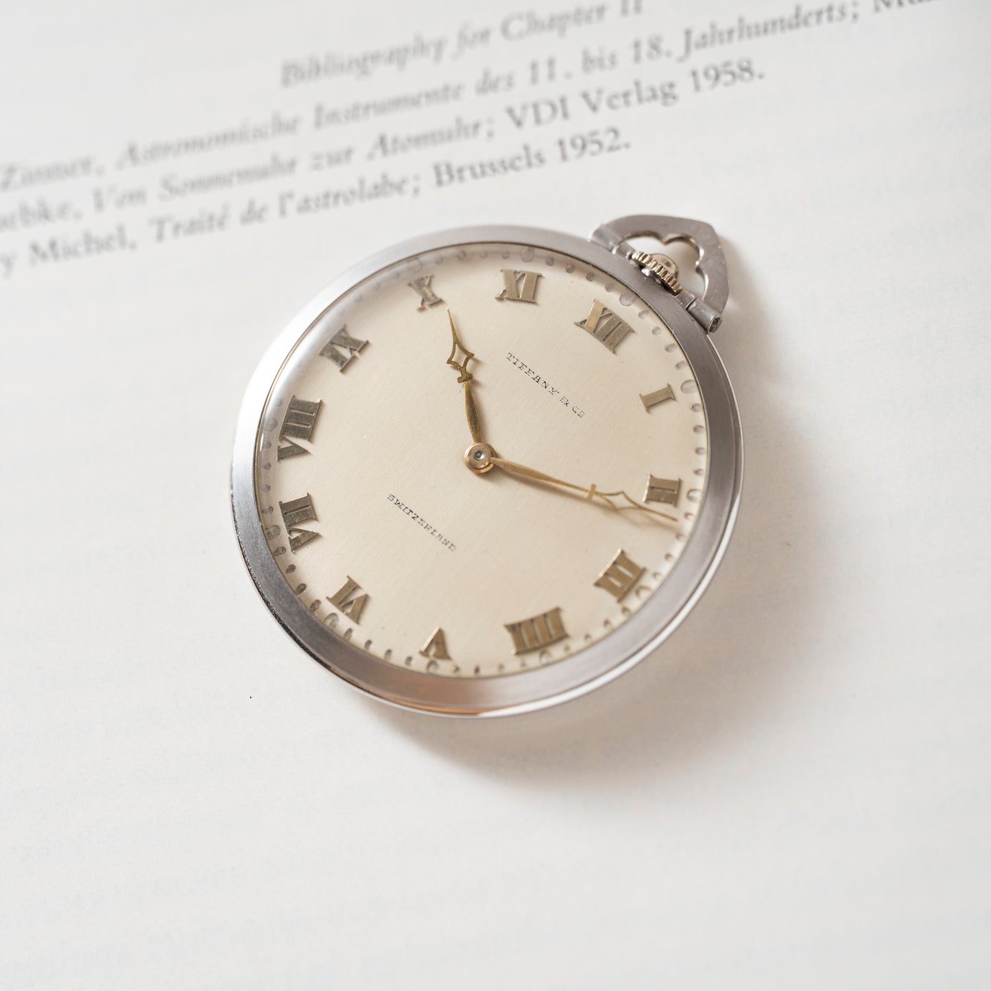 The Tiffany & Co. Platinum Pocket Watch, featuring Roman numerals and two hands, rests on an open book with its face showing about 7:23. The classic Tiffany & Co. branding is clearly visible against the printed text background.