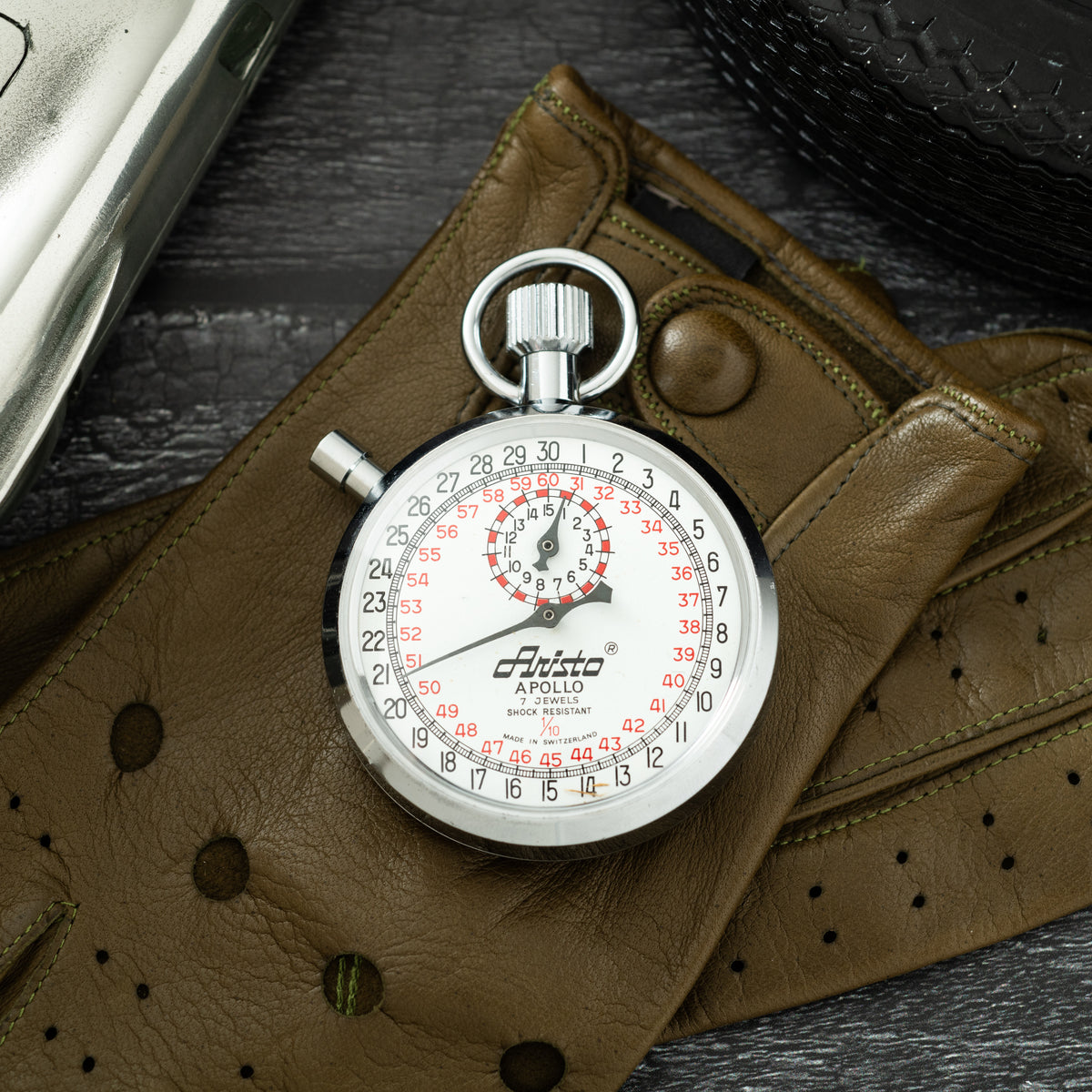 Aristo Apollo stopwatch with white dial, red numbers, and chrome case resting on tan leather pouch.